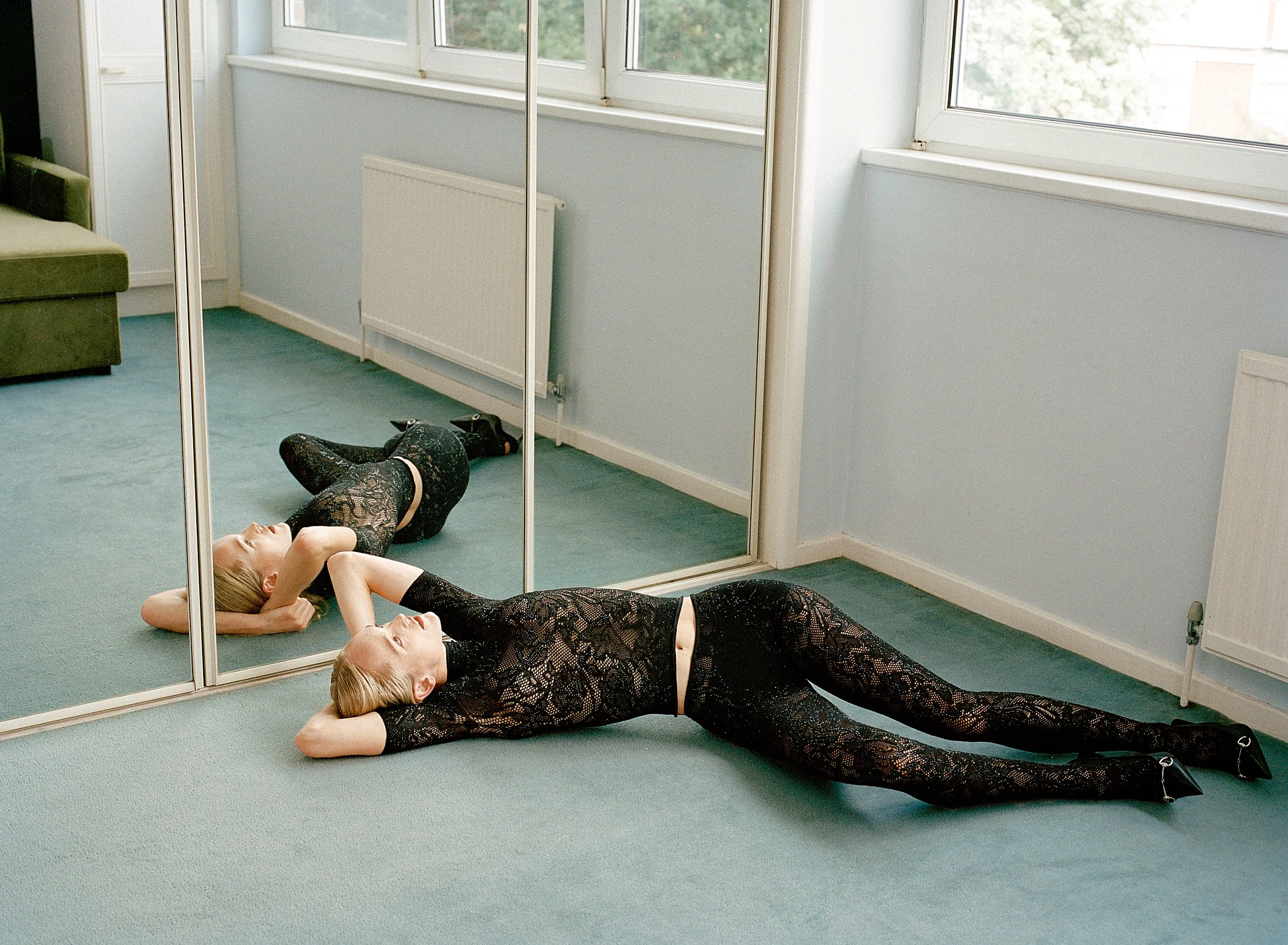 A woman in a black lace bodysuit and high heels lying on the floor in front of a mirror, stretching with her hands behind her head.