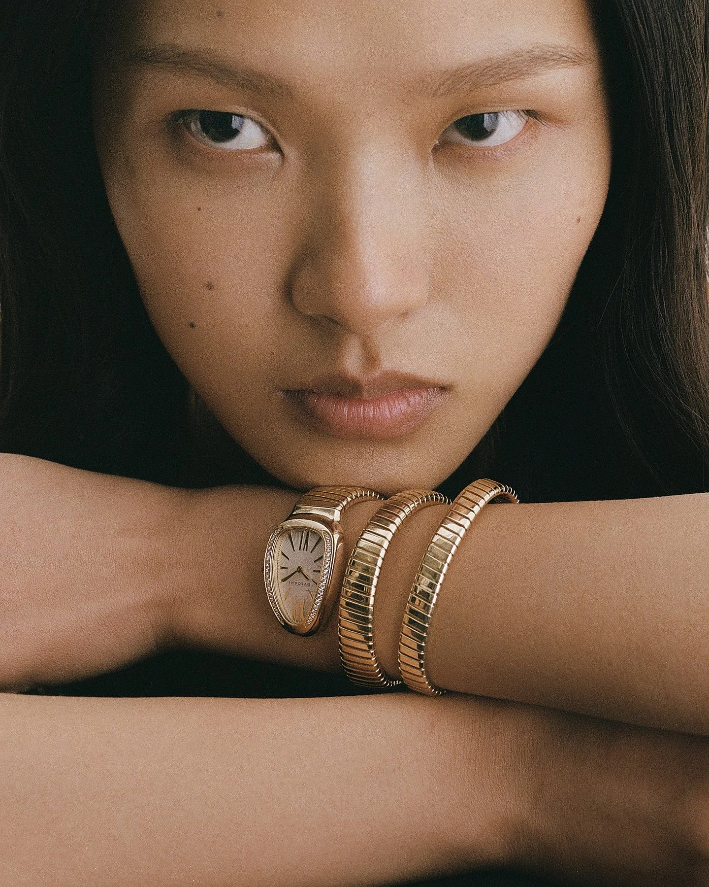 Close-up of a woman’s face with long dark hair, showing only her eyes and nose, with her arm resting on a surface. She wears a gold watch with a white face and Roman numerals and three gold bracelets on her wrist.