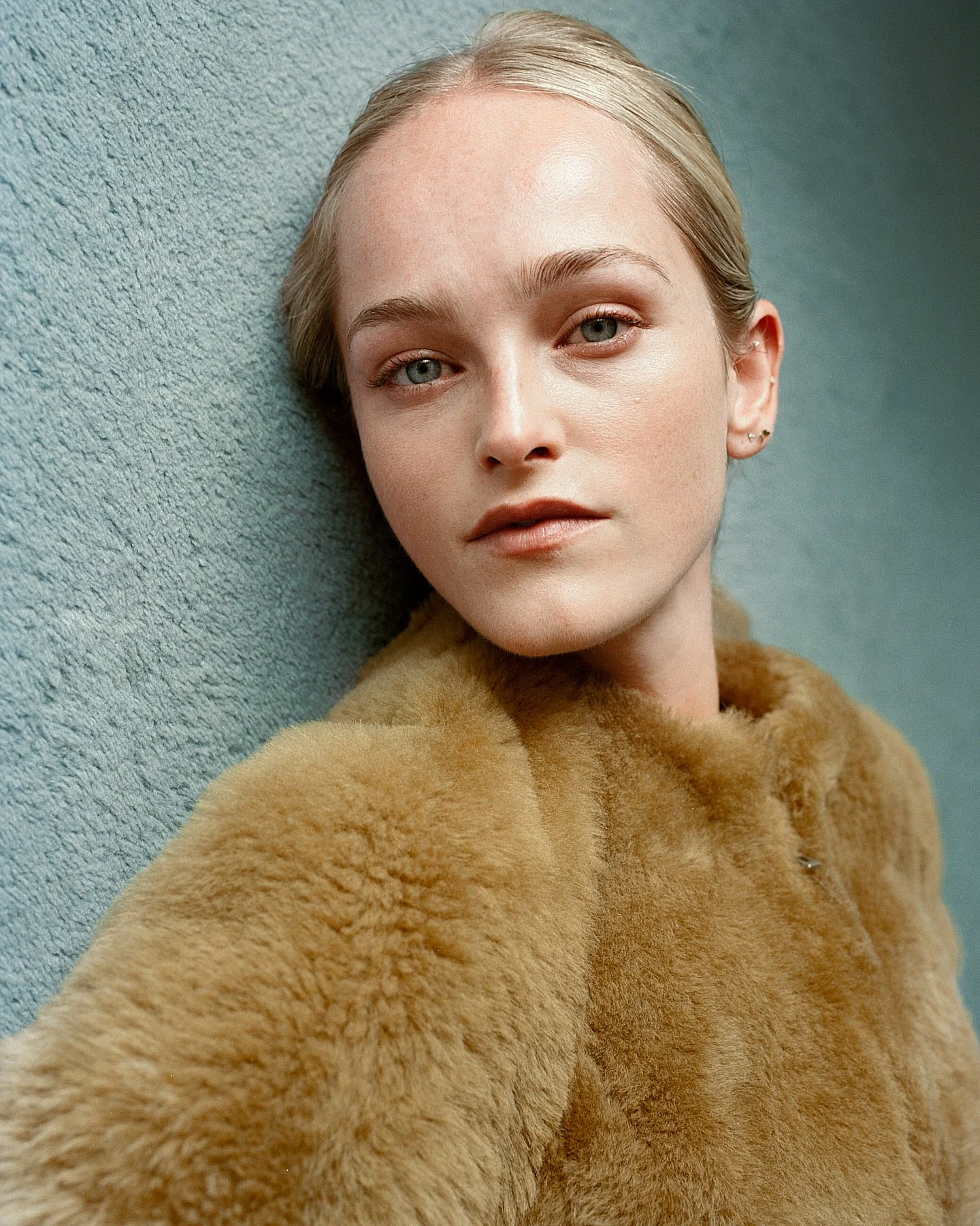 A young woman with blonde hair and blue eyes is lying on a textured blue surface, wearing a tan fur coat.