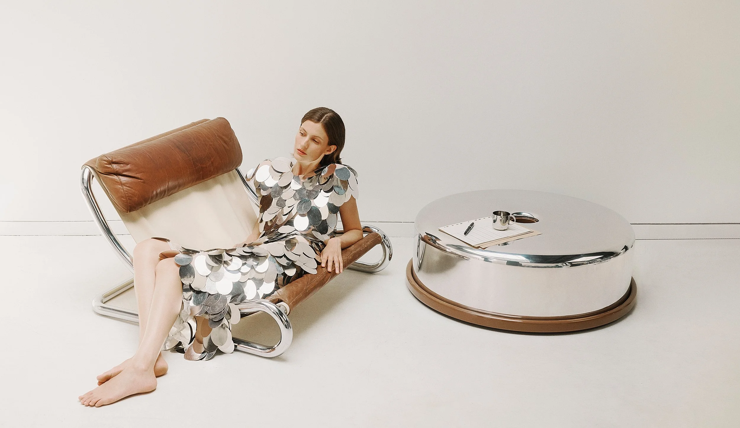 A woman in a dress made of large silver disco ball sequins sitting on a modern brown leather and chrome lounge chair in a minimalist white room with a round metallic coffee table nearby.