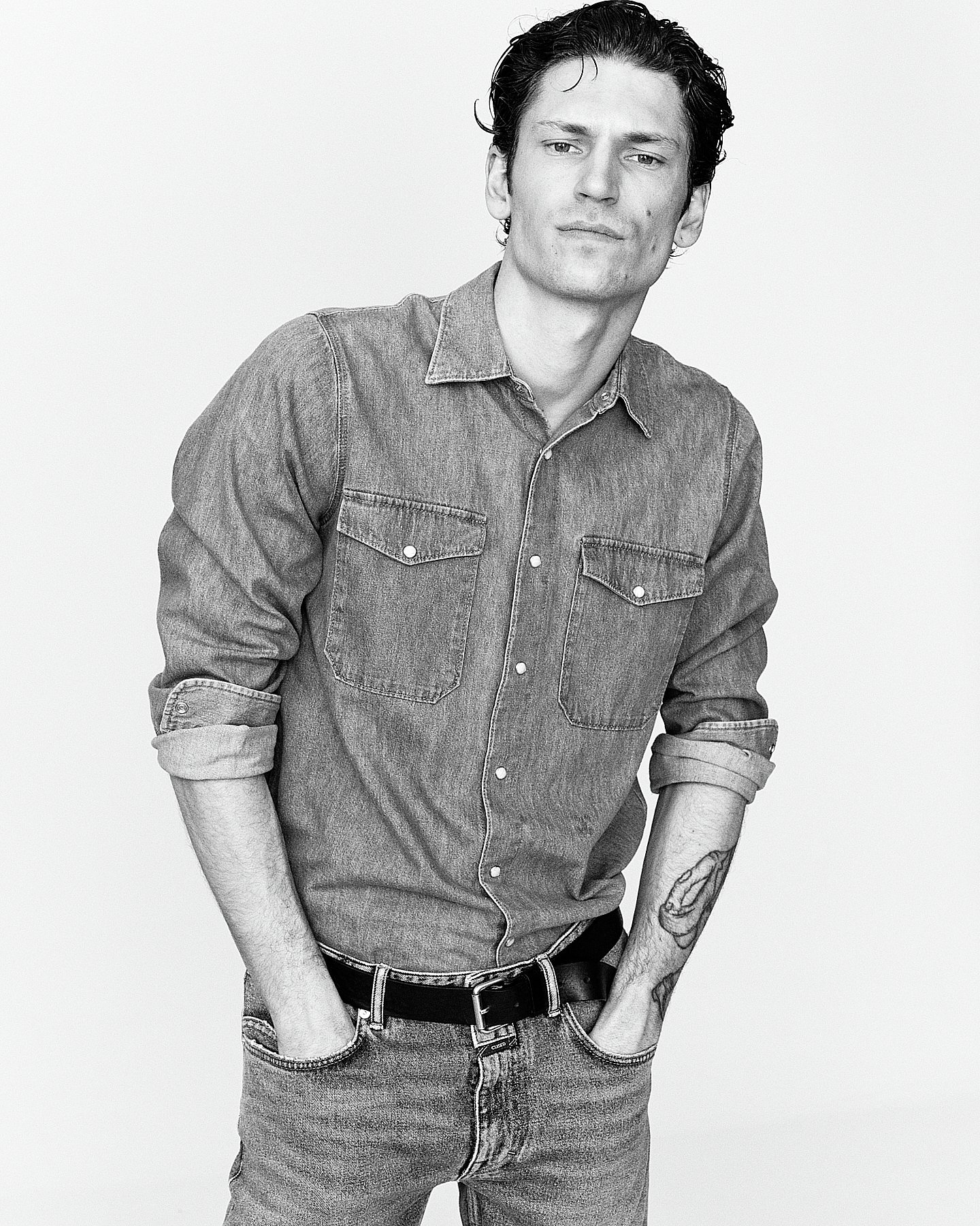 Black and white photo of a young man with dark hair, wearing a denim shirt with rolled-up sleeves, jeans, and a belt, standing confidently with hands in pockets, against a plain background.