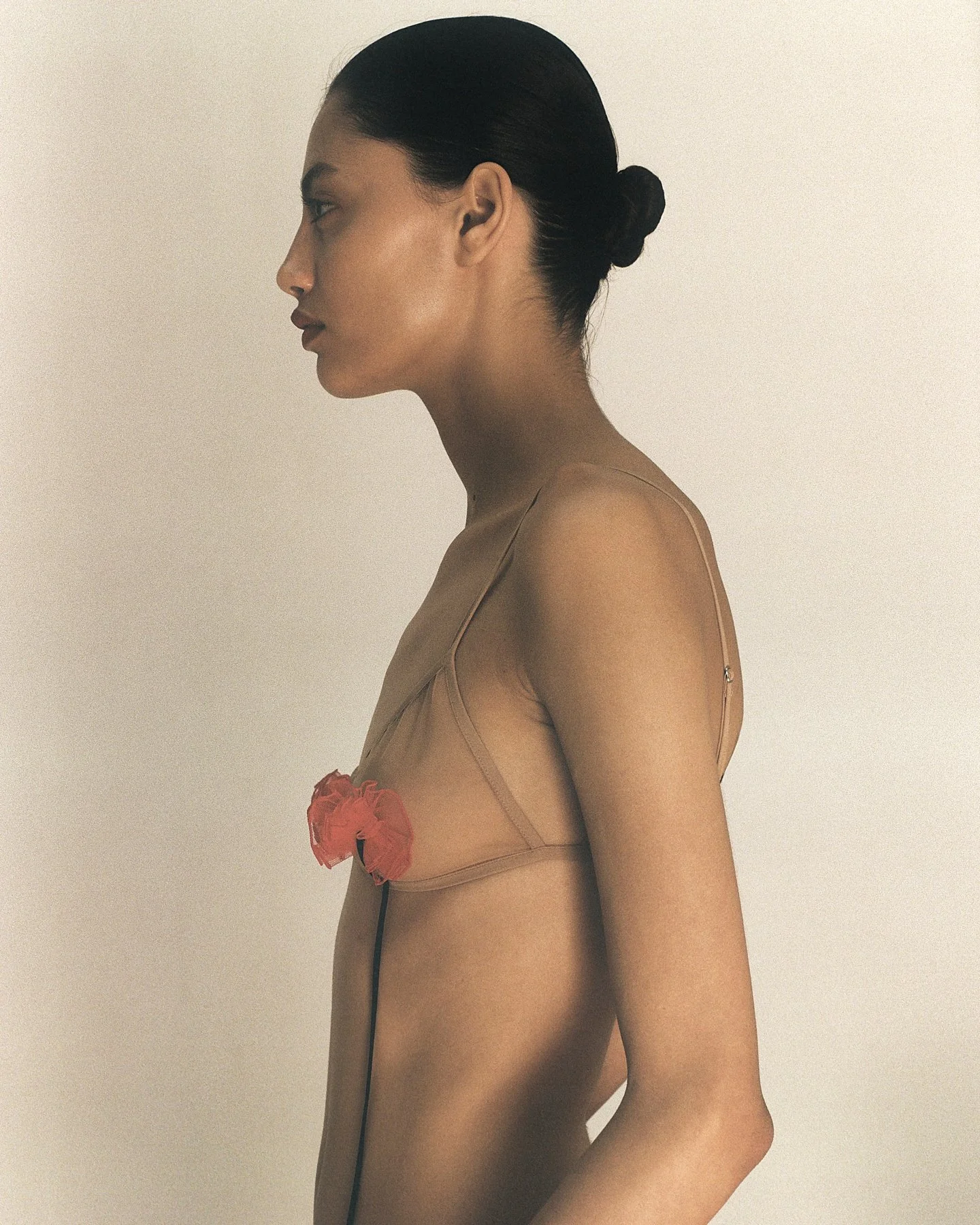 A woman with dark hair in a bun wearing sheer nude lingerie with a red flower at her side stands sideways against a plain light-colored background.