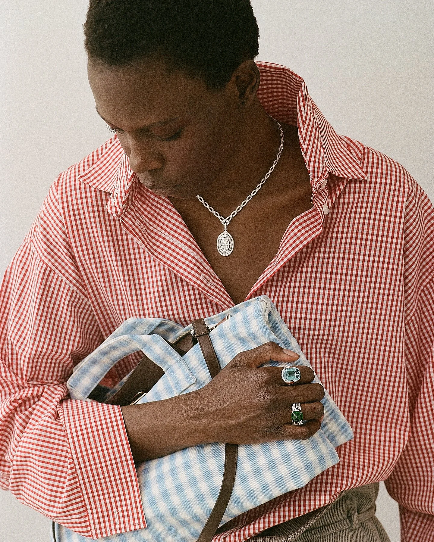 A person wearing a red and white checkered shirt, looking down and holding a blue and white checkered bag, with the focus on jewelry including rings with large stones, a silver chain necklace with an oval pendant, and holding a smartphone.