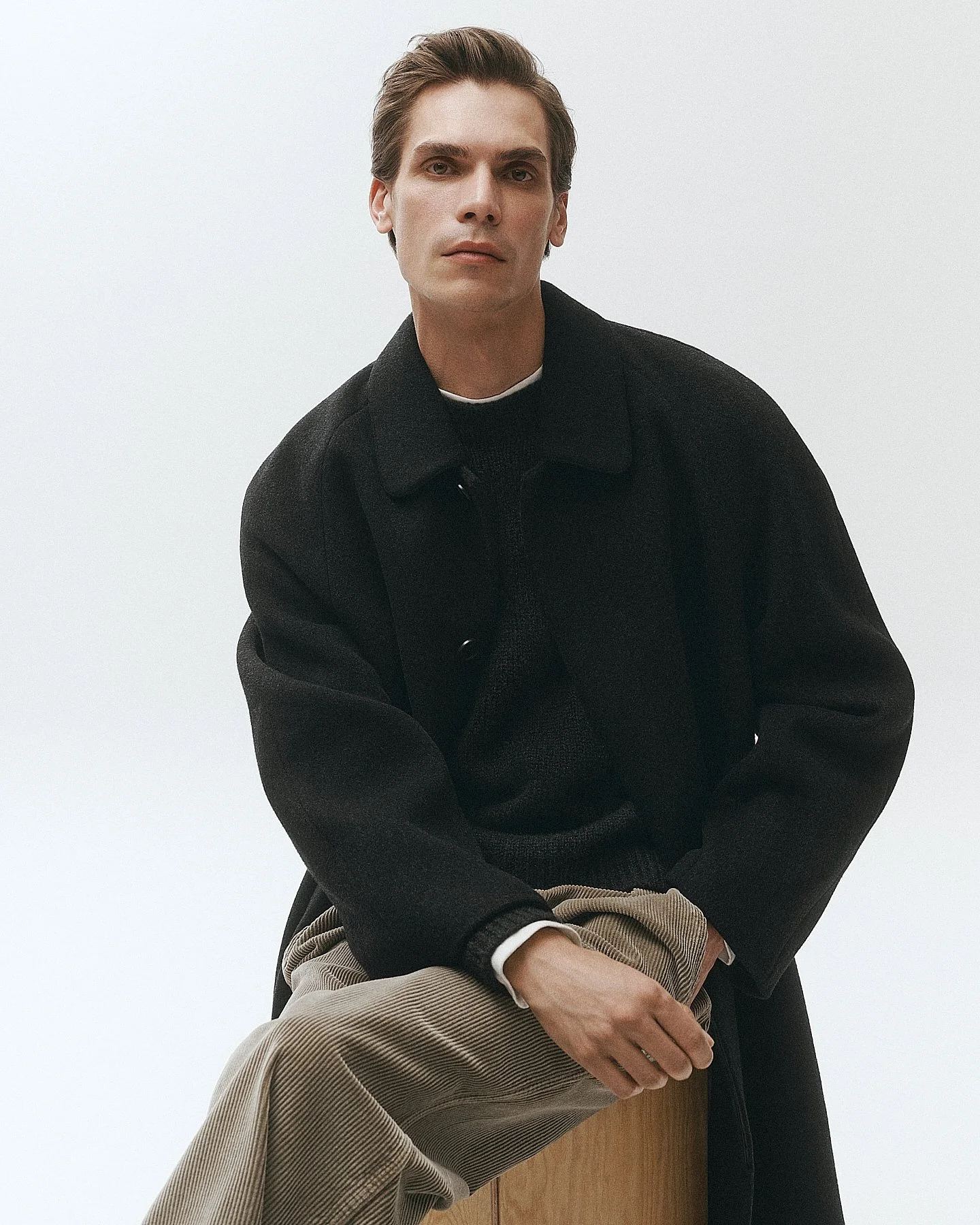 Portrait of a young man with light skin, brown hair, and blue eyes, sitting on a wooden block against a plain white background, wearing a black coat, black shirt, and beige corduroy pants.