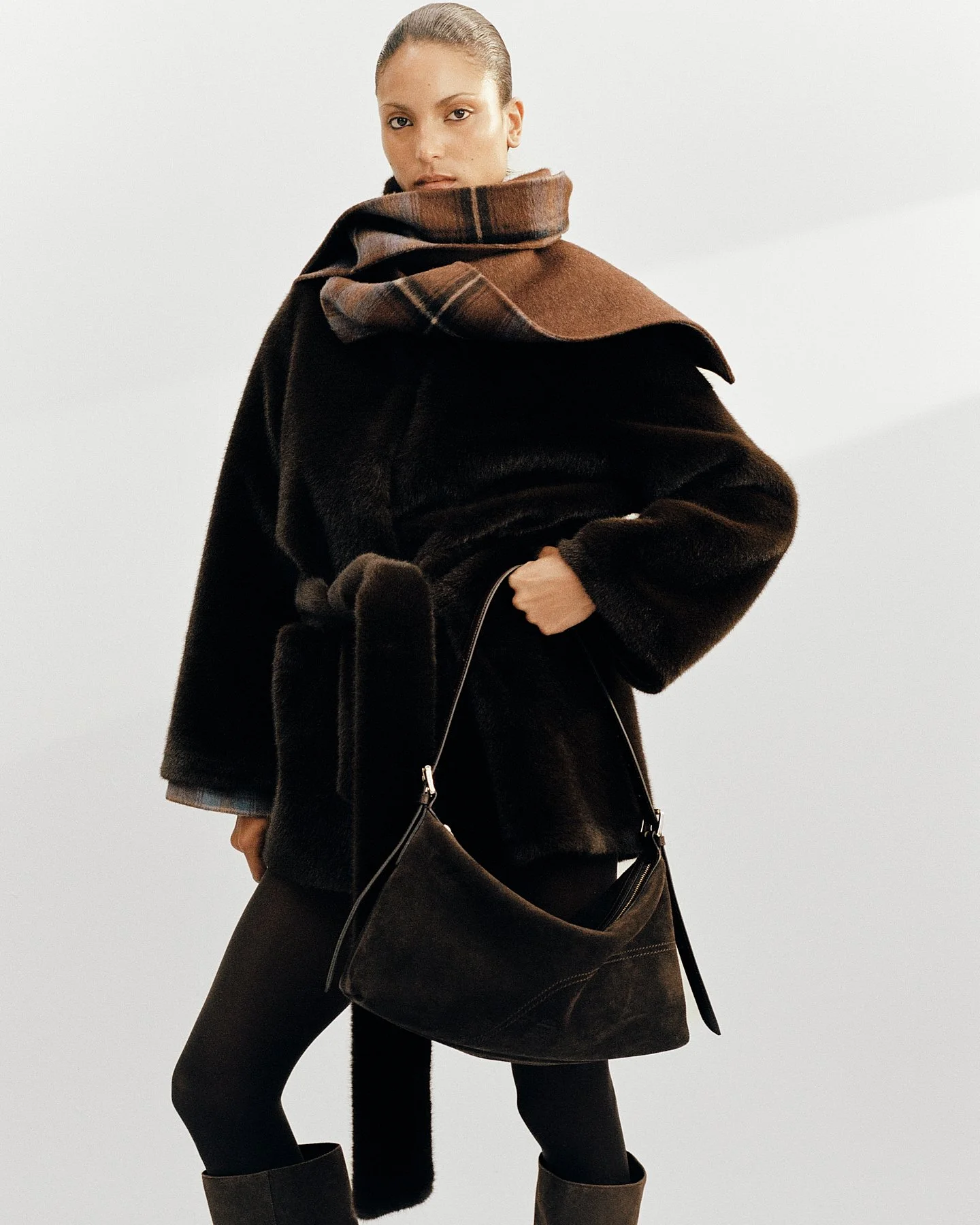 A woman wearing a black fur coat with a brown plaid scarf wrapped around her shoulders, black leggings, and boots, standing against a plain light gray background.