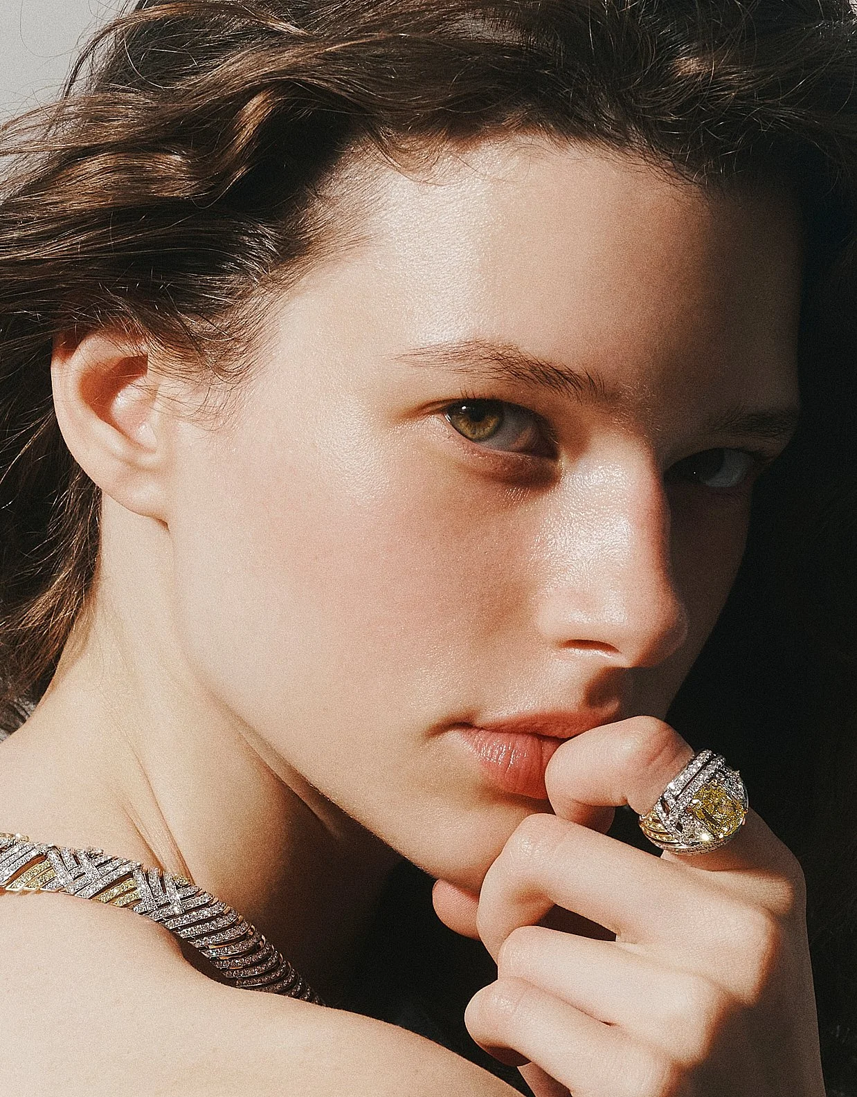 Close-up of a woman with brown hair, golden eyes, and fair skin, wearing a large diamond ring and a textured diamond necklace, with her hand near her face.