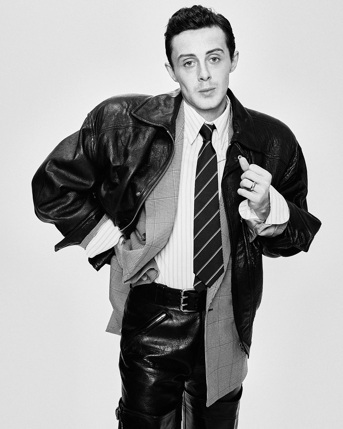 Black and white photo of a man wearing a leather jacket, striped dress shirt, and striped tie, holding a rolled-up paper in his left hand, with a confident and relaxed expression.