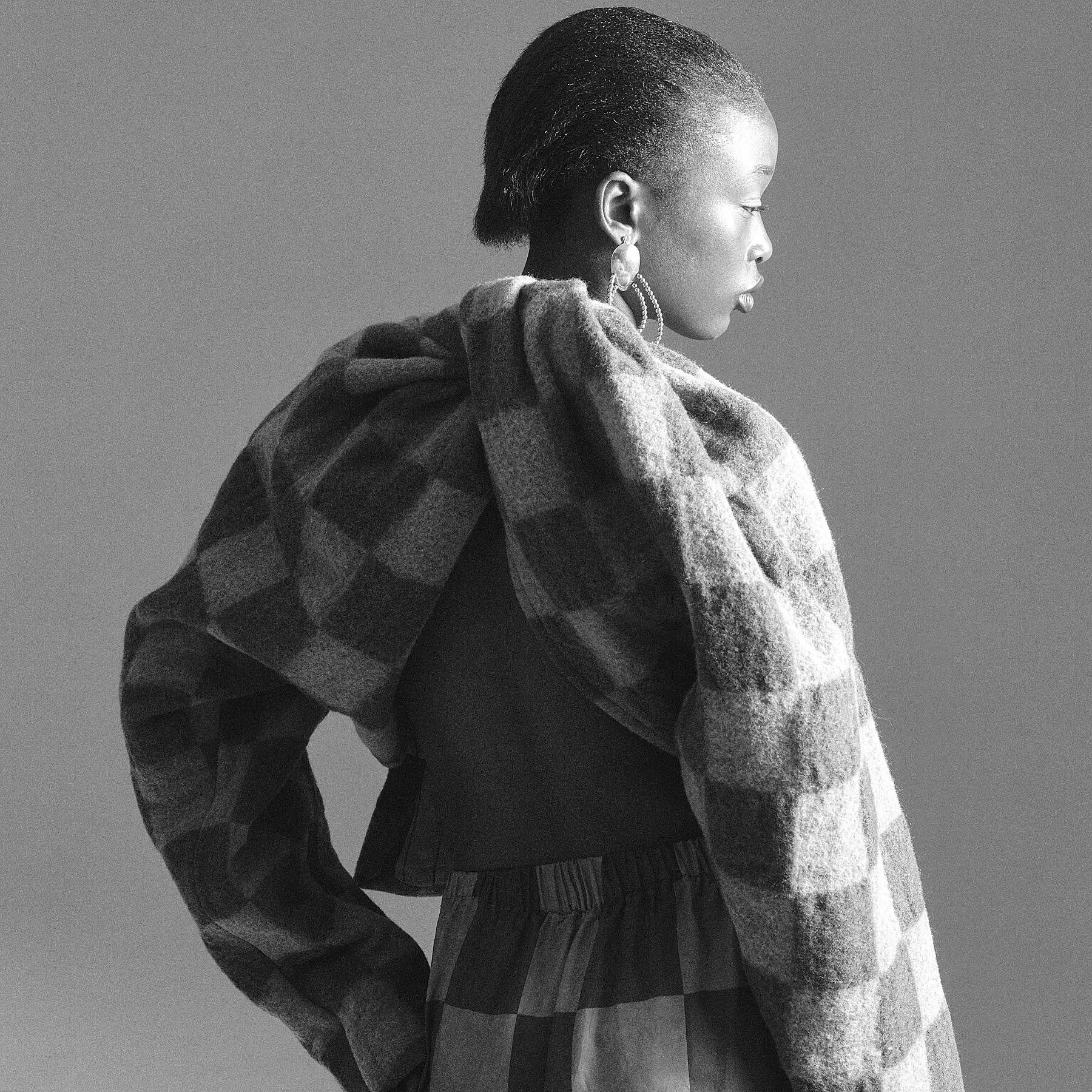 Profile of an African American woman wearing large earrings, a plaid scarf, and a puffer jacket against a plain background.