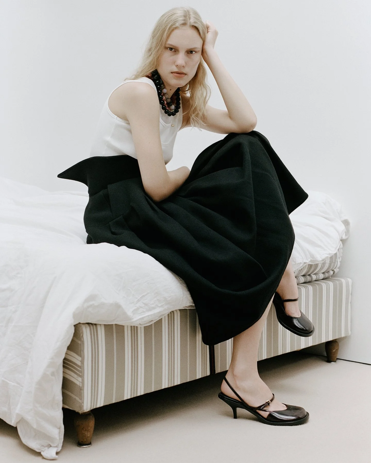 A young woman with long blonde hair, wearing a sleeveless white top, a black skirt, black high heels, and a chunky black beaded necklace, sitting on a bed with a white comforter, one leg crossed over the other, and one hand resting on her head, looki