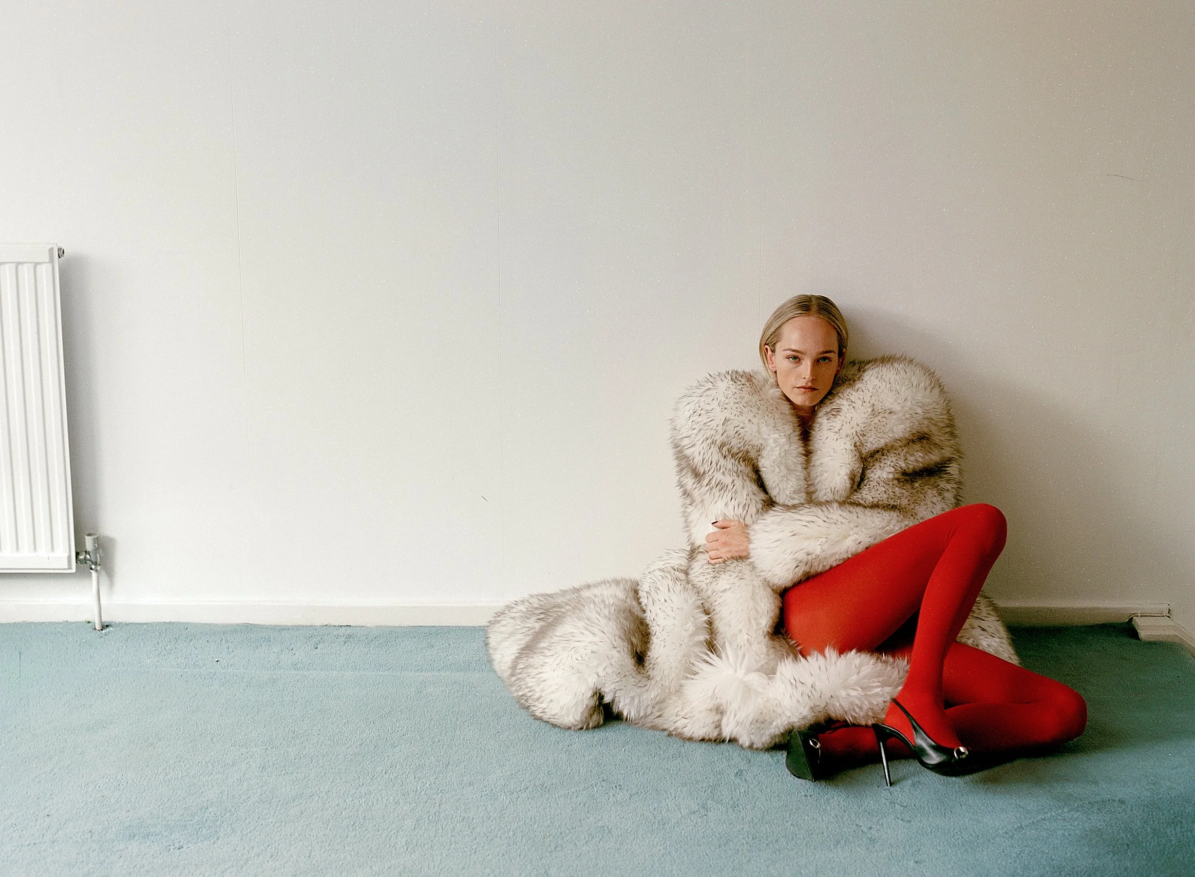 A young woman with blonde hair sitting on the floor next to a red chair with high heels, wearing a large fur coat and red pants, in a plain room with a white wall and blue-green carpet.