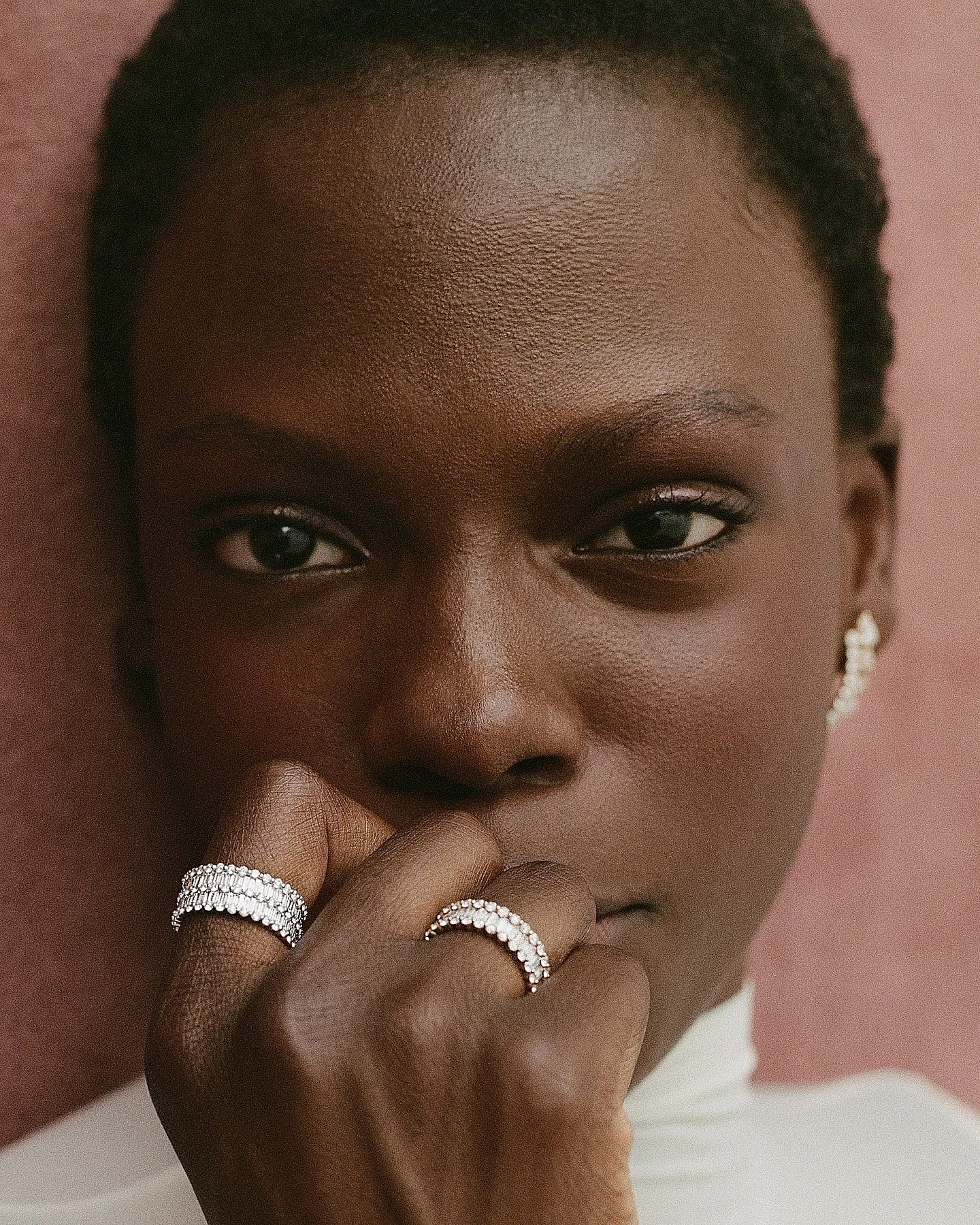 A close-up of a woman with dark skin, wearing rings on her fingers and an earring, with her hand near her chin and her eyes looking into the camera against a pink background.