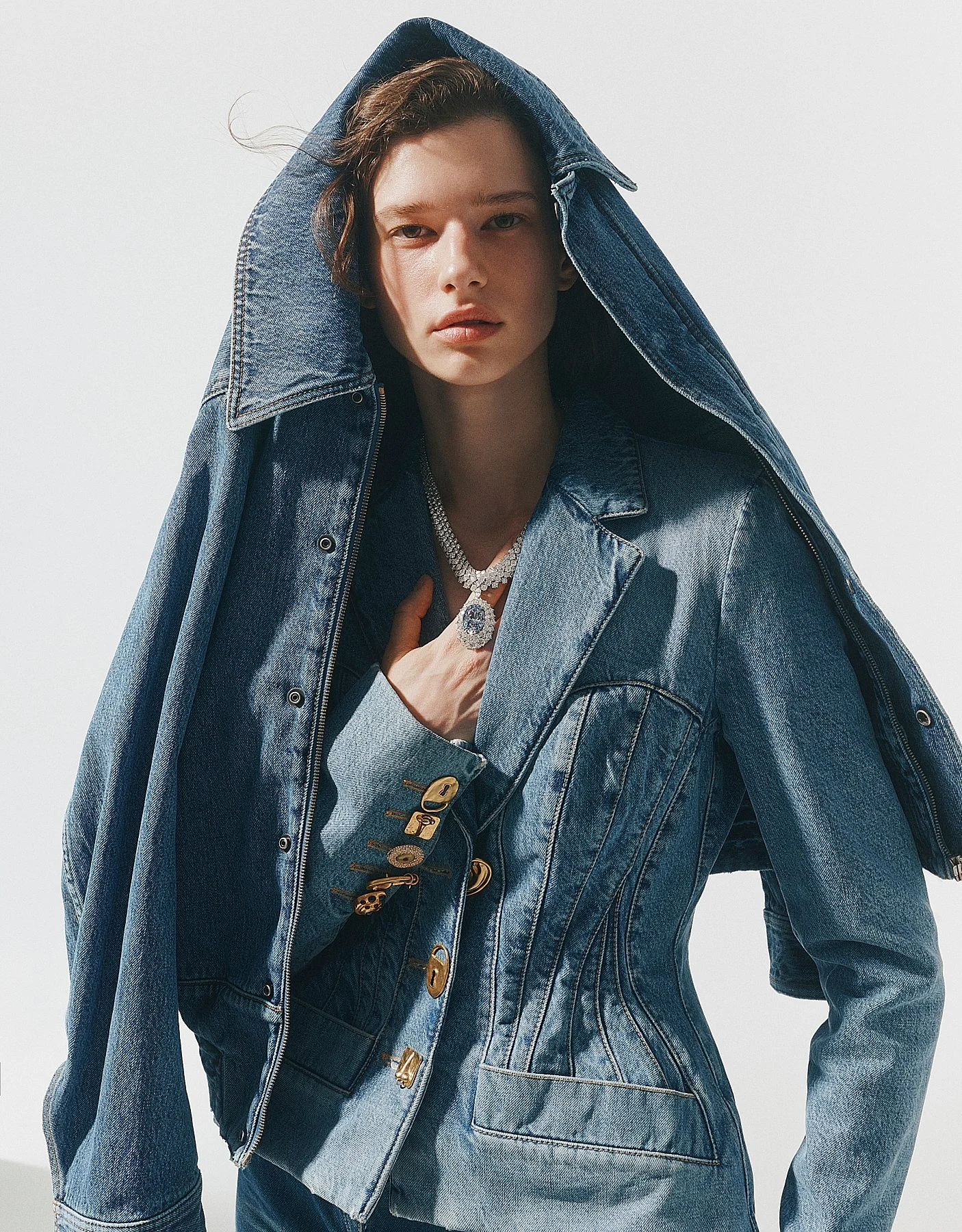 A woman wearing a blue denim jacket with the hood up, accessorized with jewelry, against a plain light background.