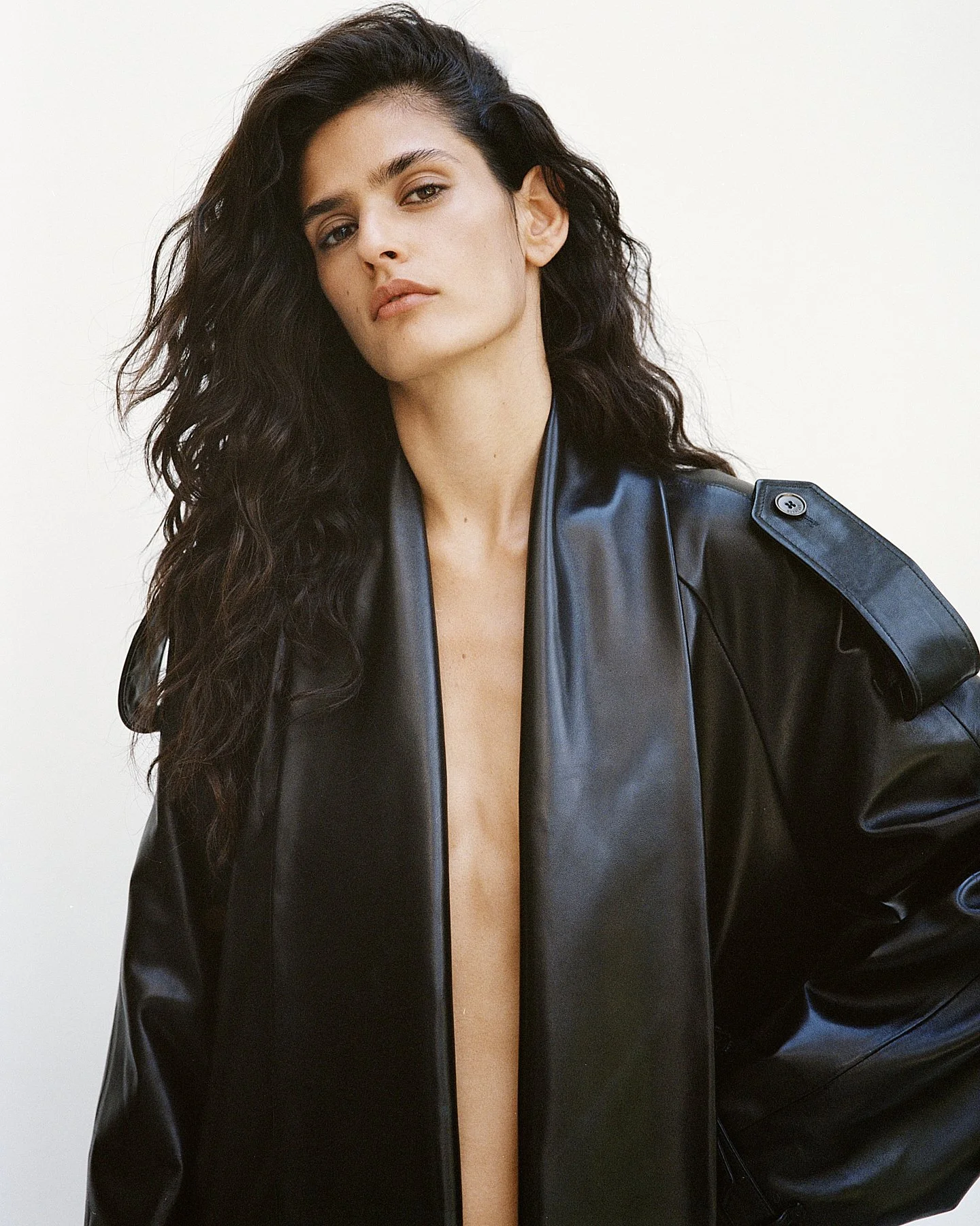 A woman with long dark wavy hair, light skin, and a serious expression wearing a black leather jacket open at the front, revealing her bare chest, standing against a plain light background.