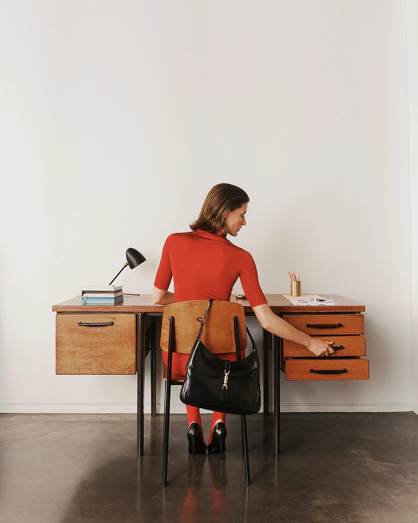 Woman in a red dress sitting at a wooden desk, reaching into a drawer, with a black handbag hanging on her chair, and a lamp, books, and a gold container with pencils on the desk.