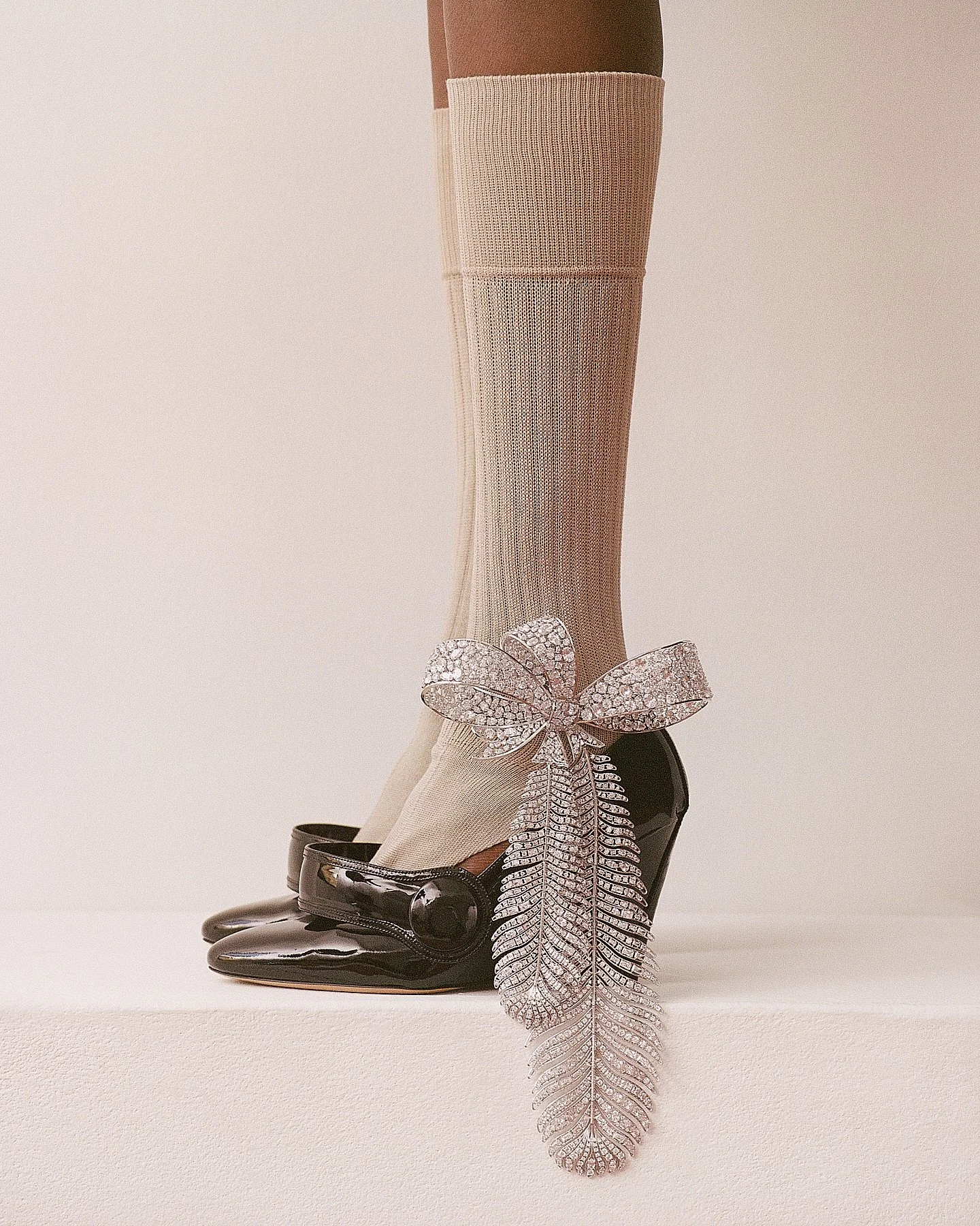 Display of a mannequin leg wearing beige fishnet stocking with a decorative glittering butterfly and leaf charm, black patent leather Mary Jane shoes, and a black high heel, against a light neutral background.