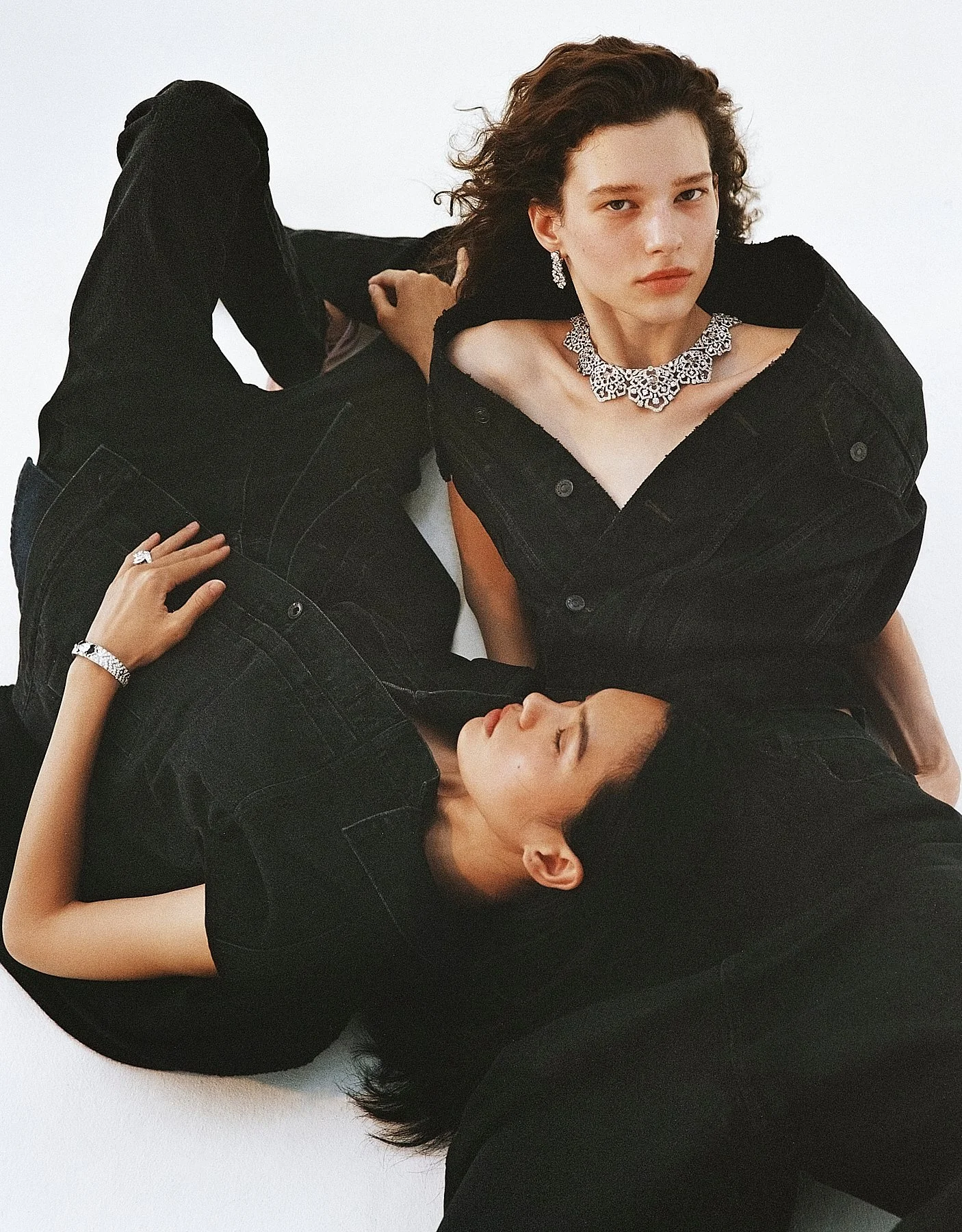 Three women dressed in black clothing and jewelry, lying on a white background, with one woman looking directly at the camera while the other two women are resting with their eyes closed.