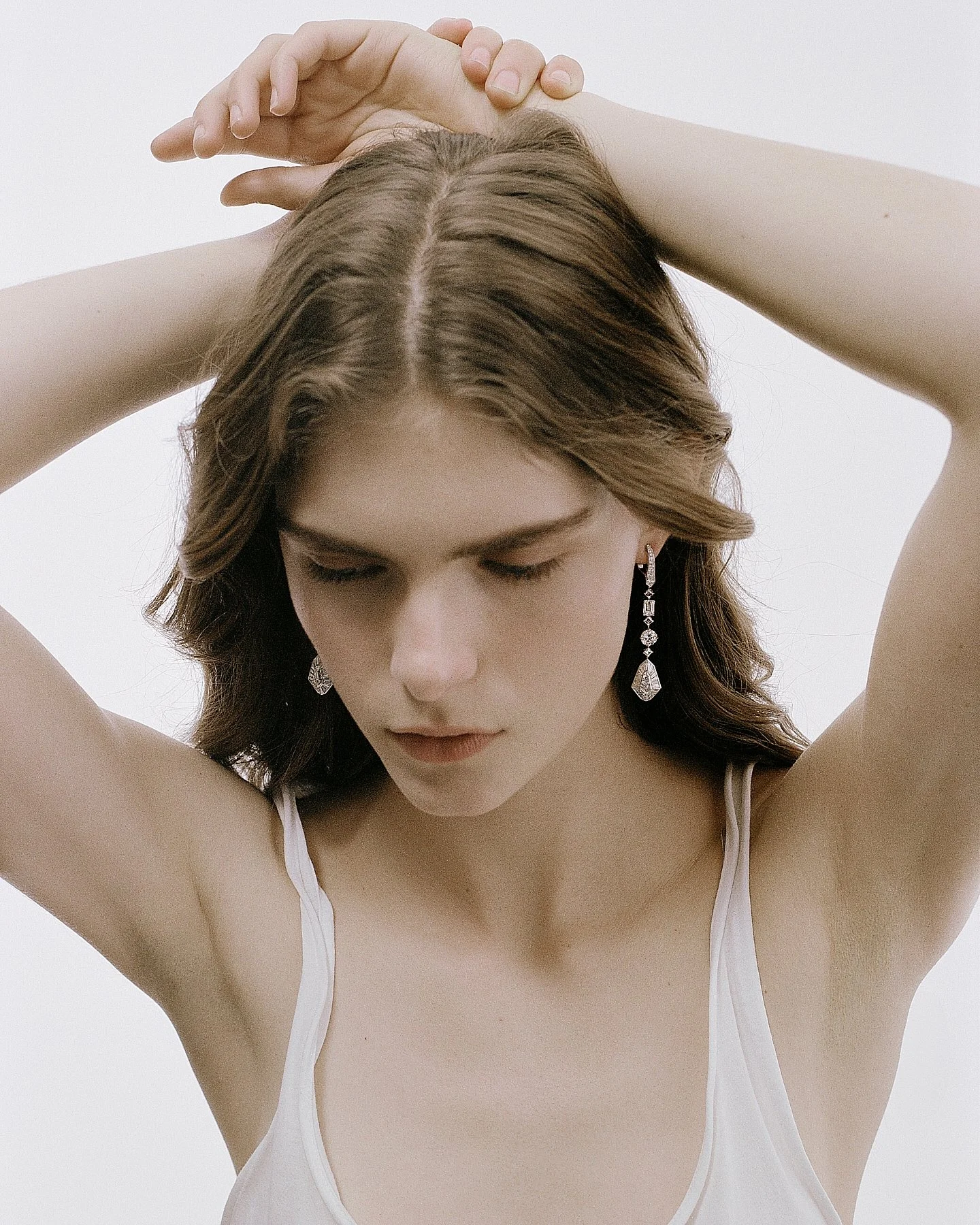 A woman with wavy brown hair wearing dangling earrings and a white spaghetti strap top, with her arms raised and hands behind her head, against a plain white background.