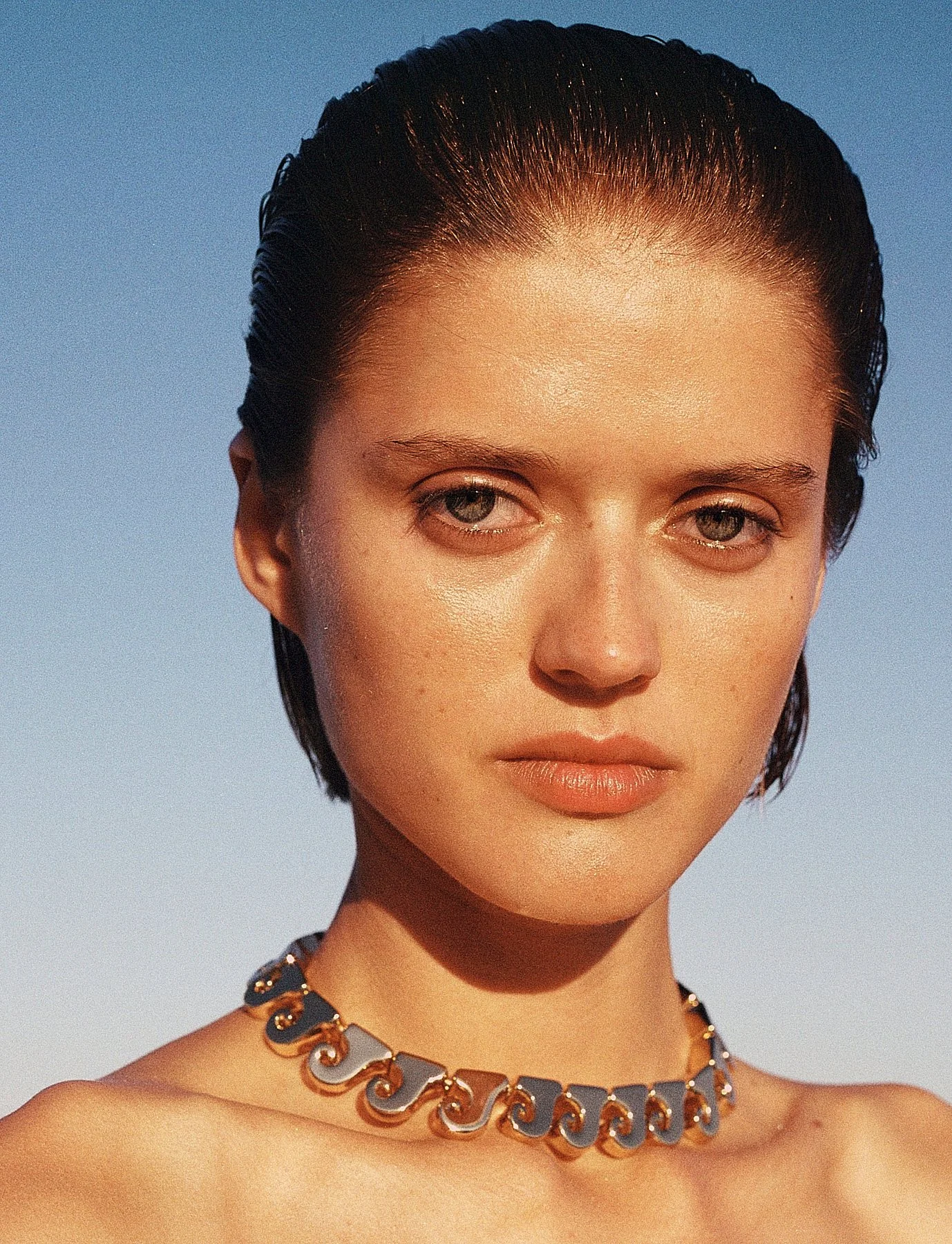 Close-up of a woman with short, dark, wet-looking hair, fair skin, and neutral makeup, wearing a gold-colored jewelry necklace. The background shows a clear blue sky.