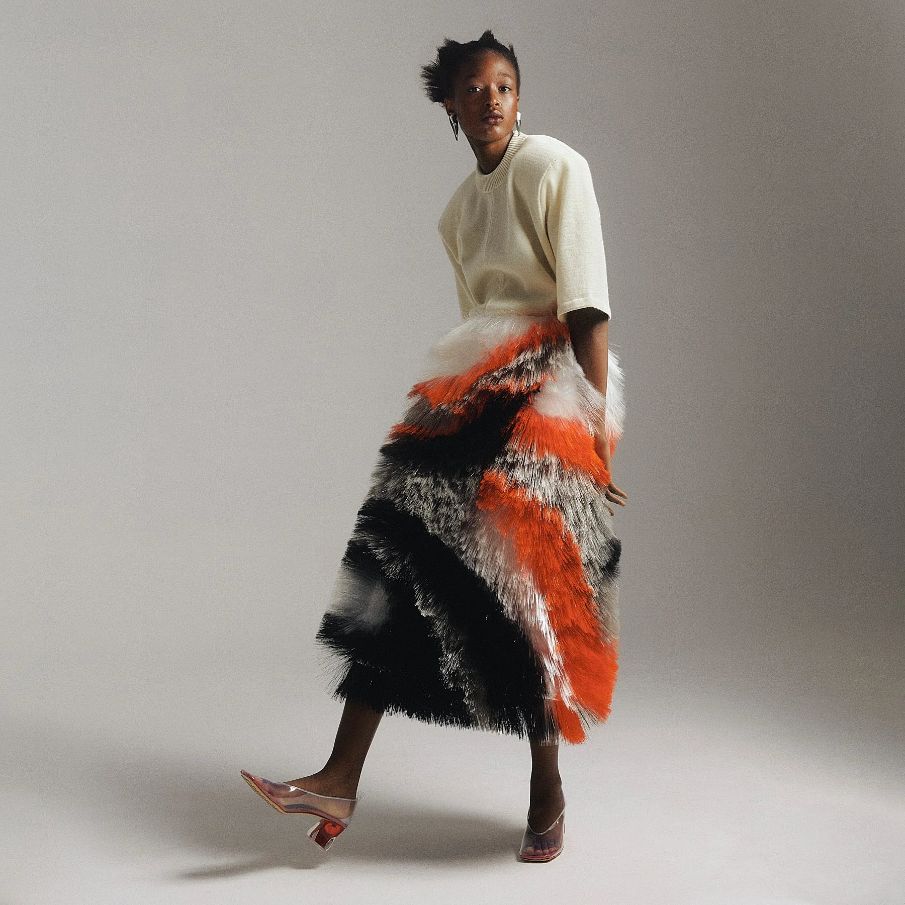 Fashion model walking in a studio wearing a cream oversized sweater, a colorful feathered skirt with orange, black, white, and pink feathers, high heels with floral pattern, and statement earrings.