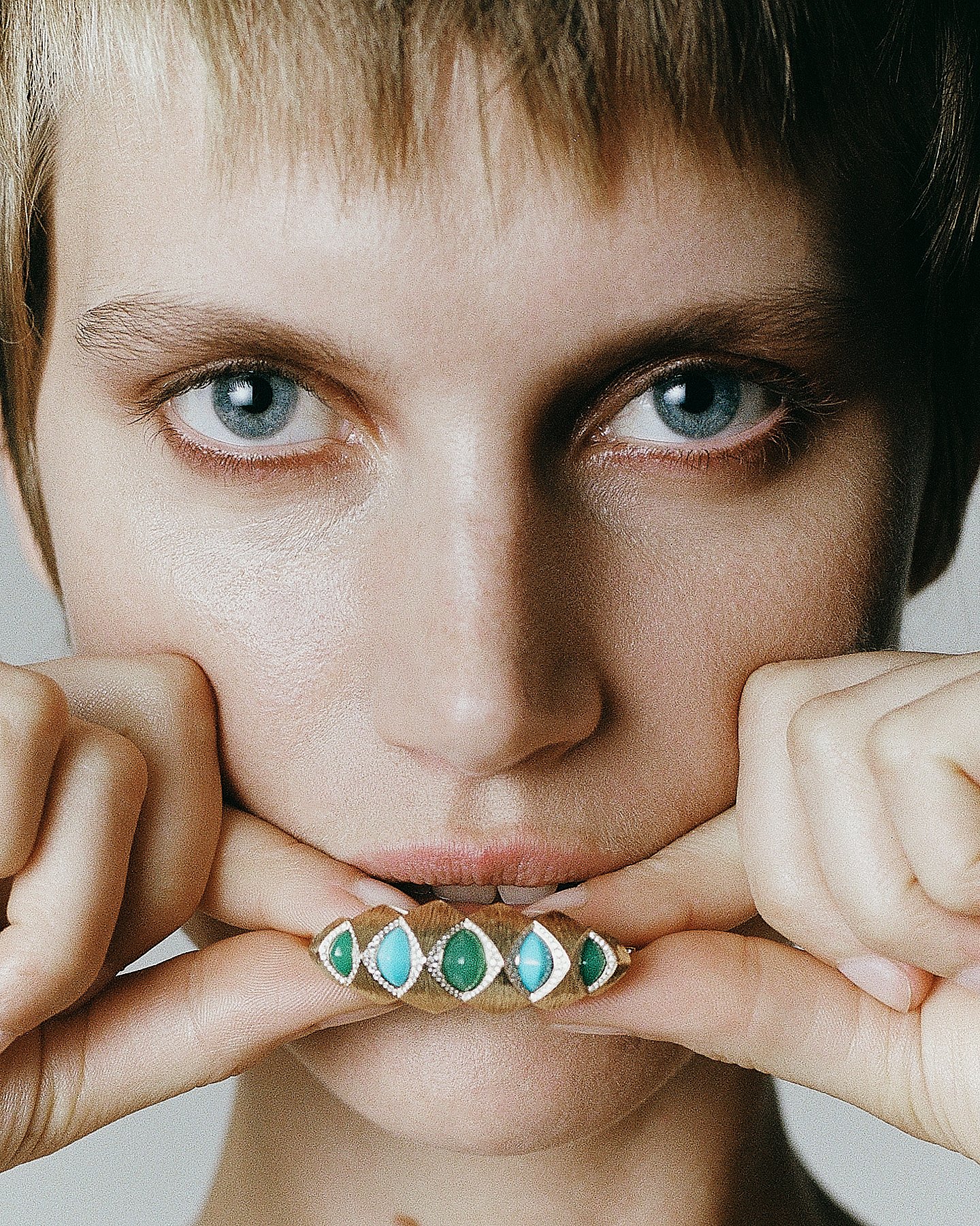 Close-up shot of a young woman with blue eyes holding a ring with green and blue gemstones near her lips, with her fingers framing her face.