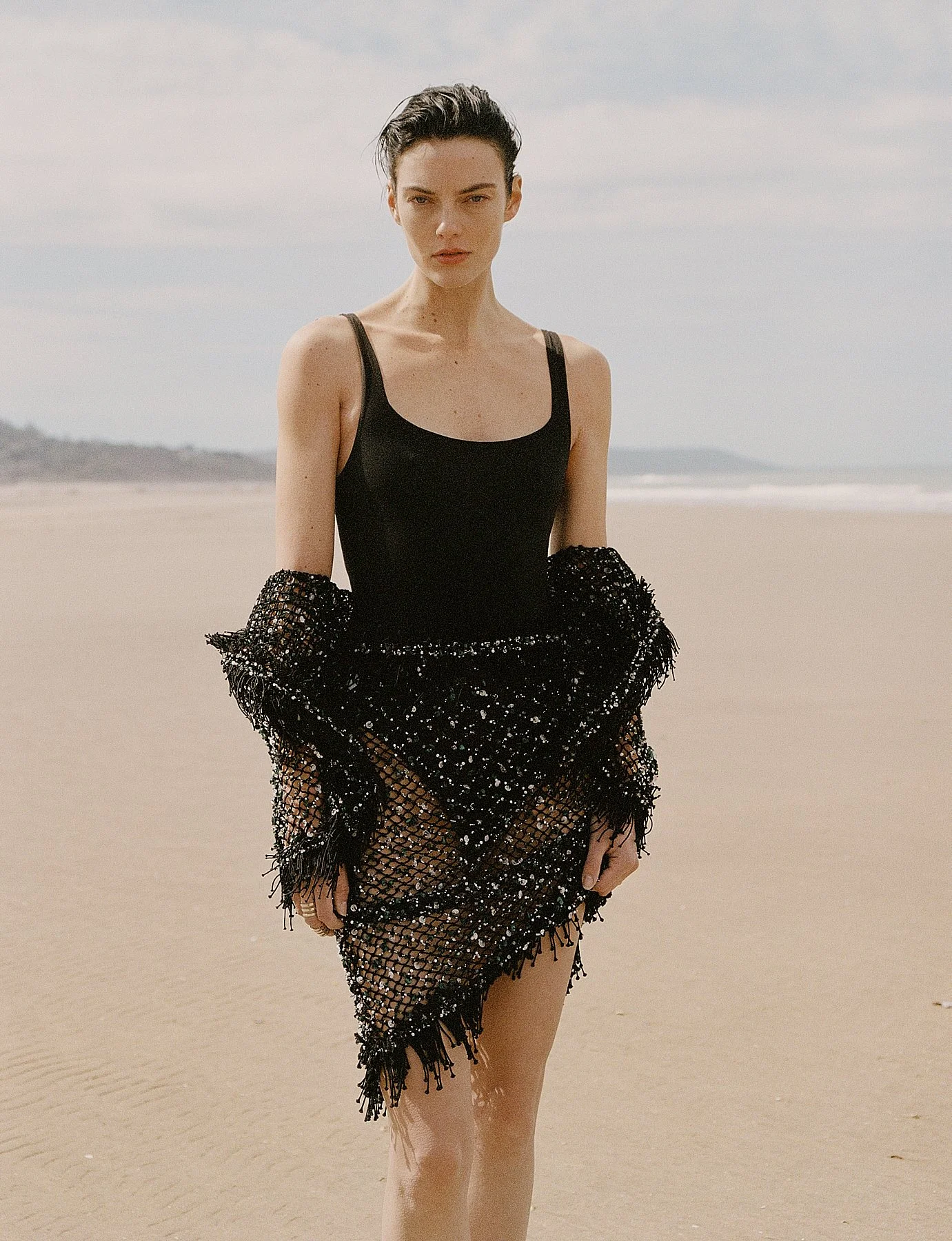 A woman standing on the beach wearing a black tank top and a black, sequin-covered skirt with fringes, holding a matching black shawl.