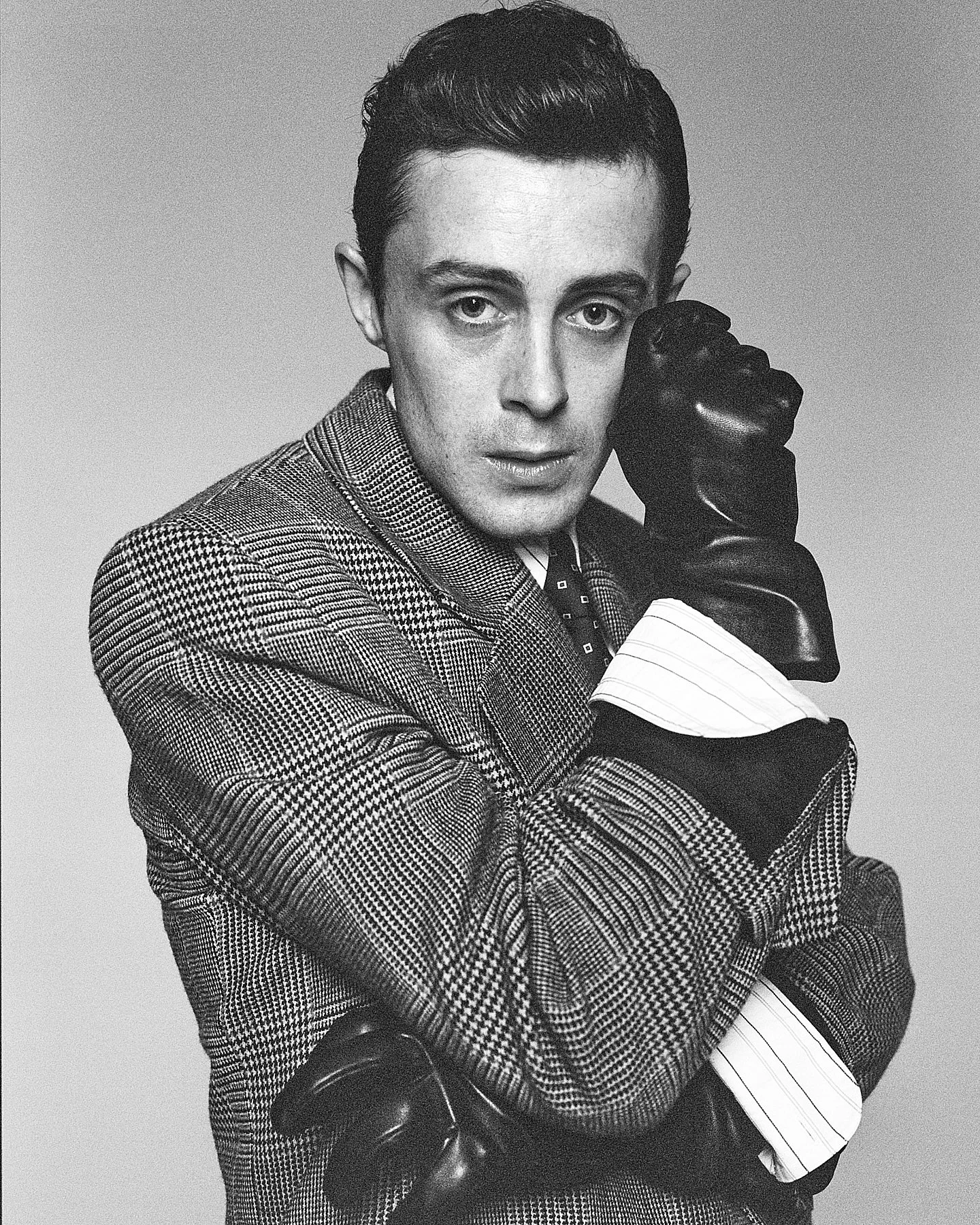 Black-and-white photo of a person with short, styled hair wearing a patterned suit, with one arm crossed and the other hand resting on their face, which is covered by a black glove.
