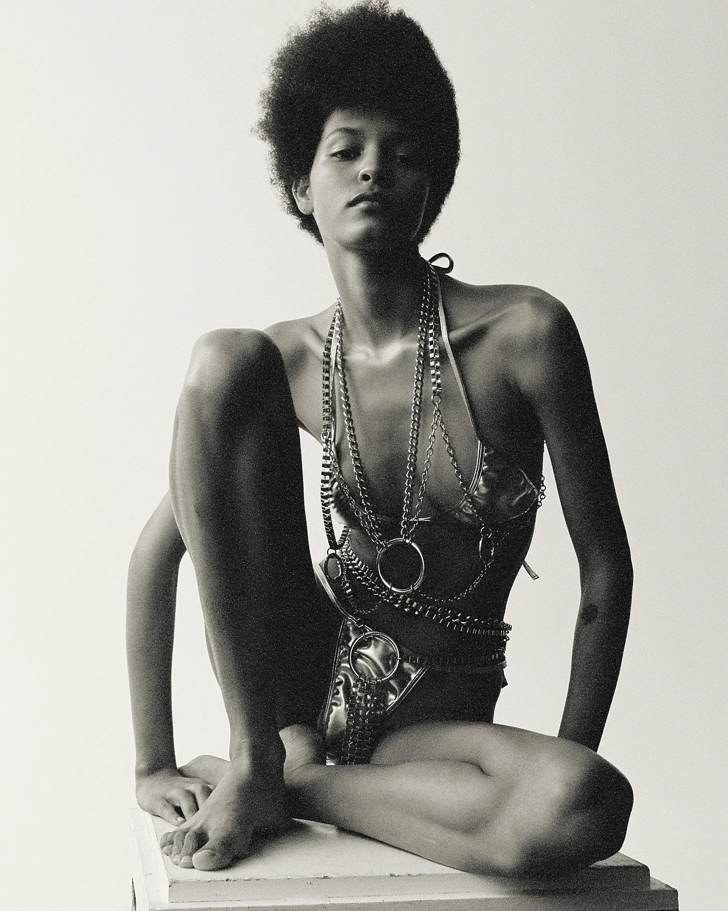 Black-and-white photograph of a mannequin with an afro hairstyle, adorned with many layered chains and jewelry, seated with one knee up and one hand resting on the ground, against a plain background.