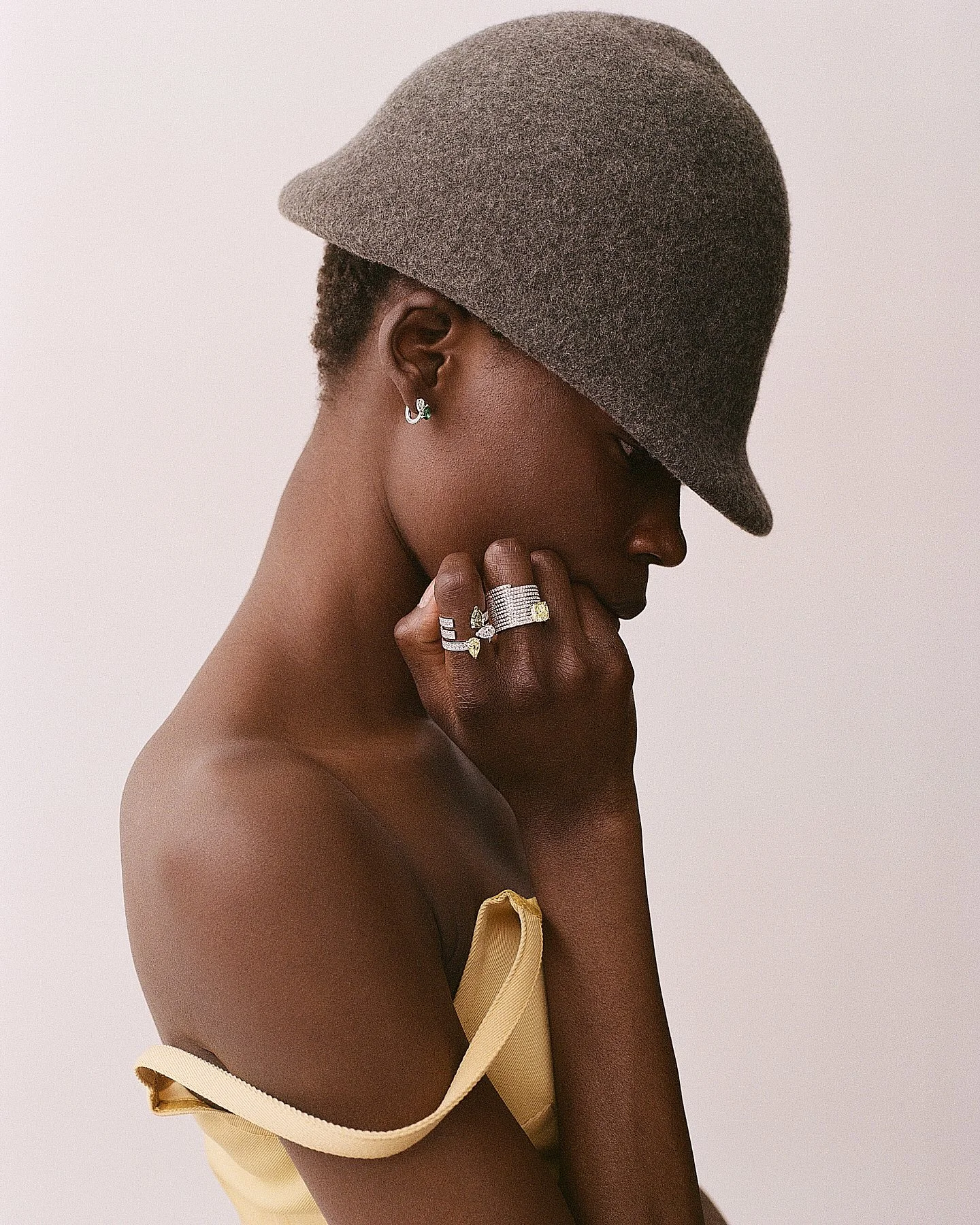 A woman with dark skin wearing a gray hat, silver jewelry including earrings and rings, and a beige strapless top against a plain background. She is posed in profile with her hand near her face.