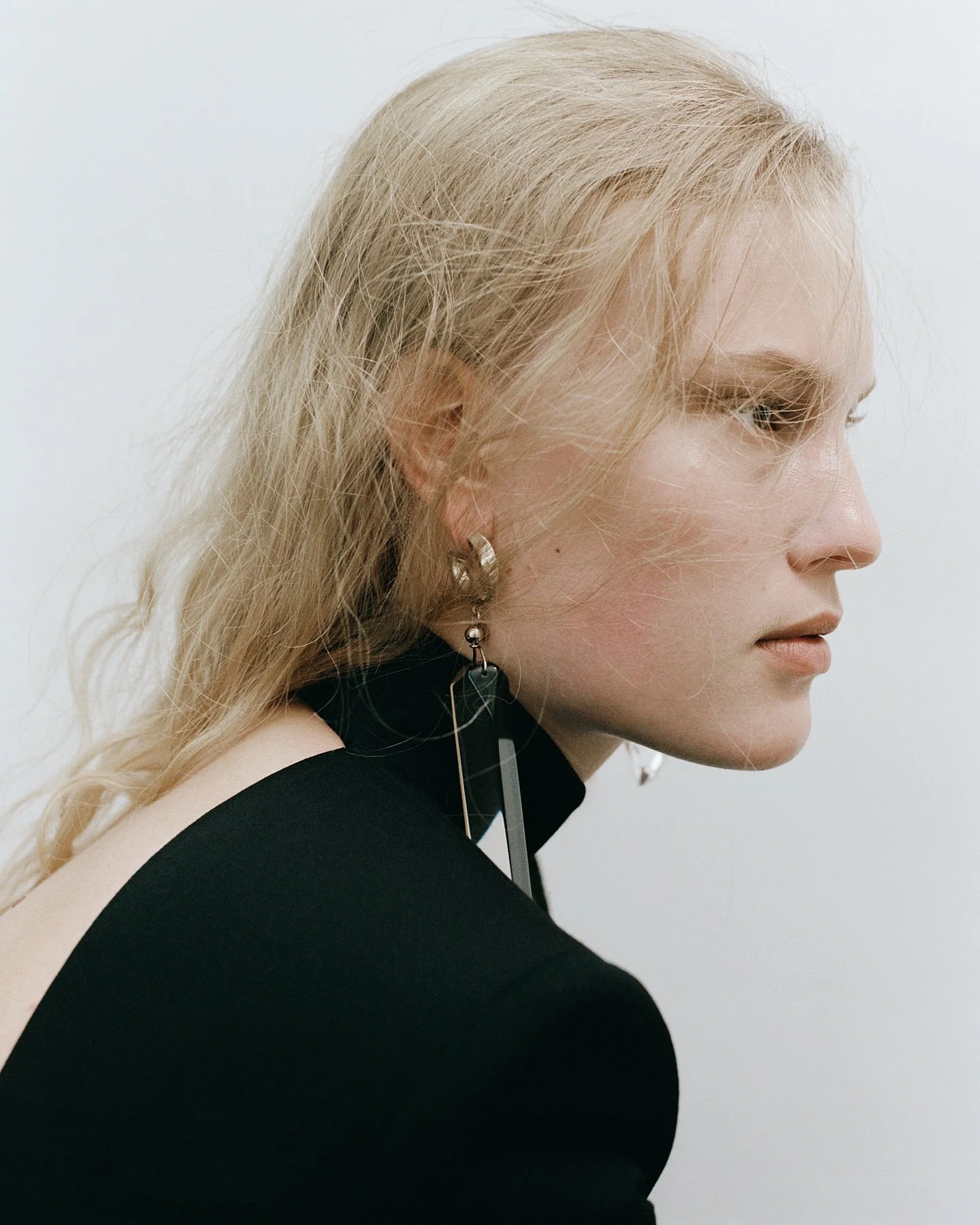 Side profile of a woman with blonde, messy hair, wearing a black top with a cutout on the shoulder, silver hoop earrings, and earphones, against a plain white background.