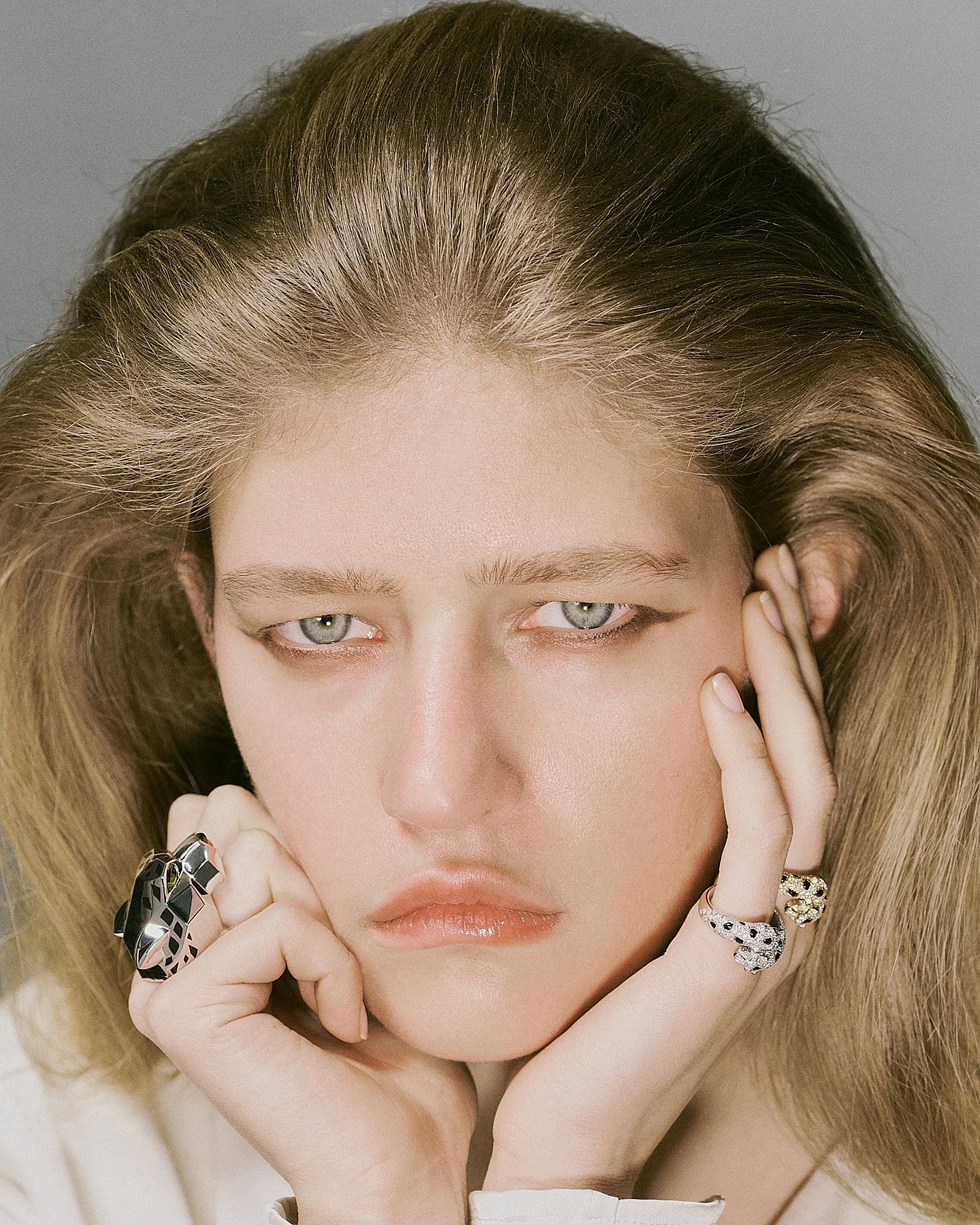 Close-up of a woman with blonde hair and light blue eyes, touching her face with her right hand, wearing rings.