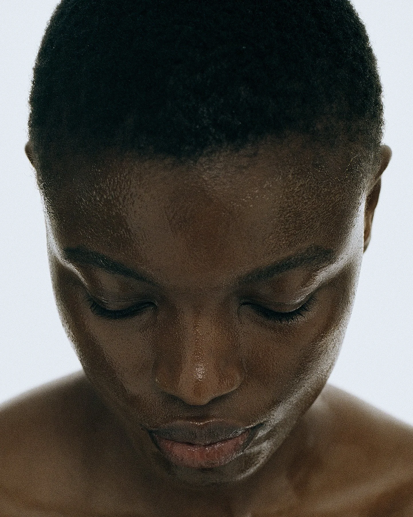 Close-up of a person's face with eyes closed and head slightly tilted downward, showing short curly hair and smooth skin.