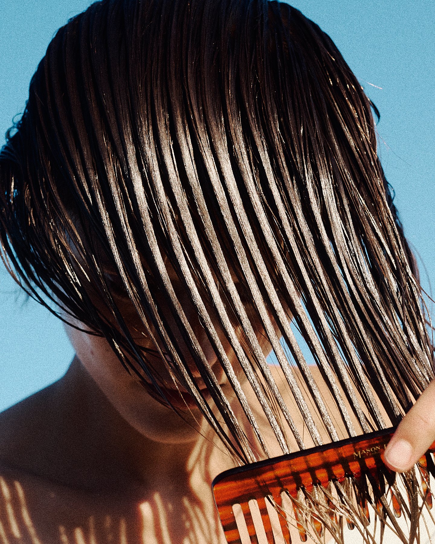 A person with wet, straight brown hair partially covering their face, holding a hair comb against their hair with a blue background.