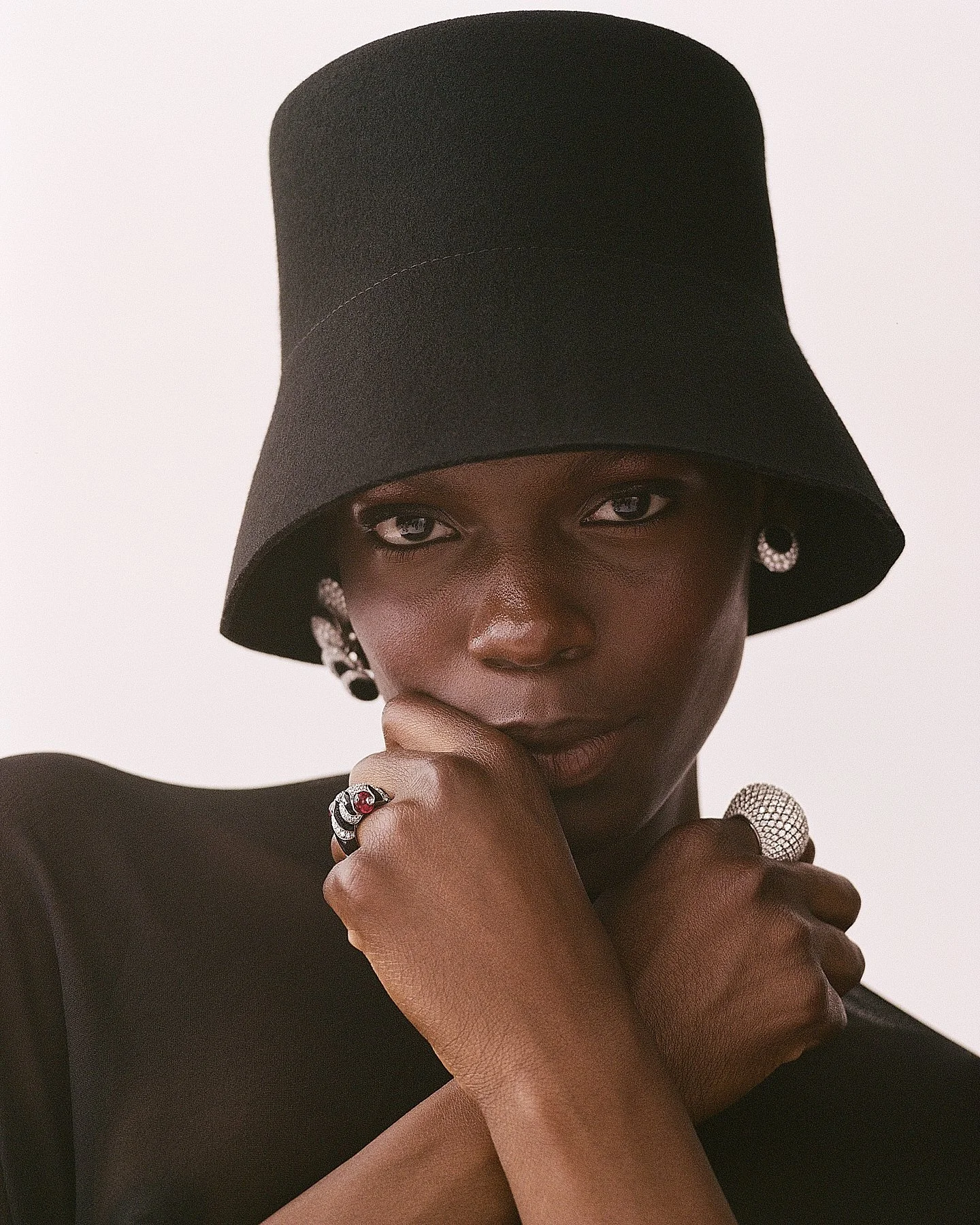 A woman with dark skin wearing a large black hat, black dress, and silver jewelry, posing with her hand near her face against a plain white background.