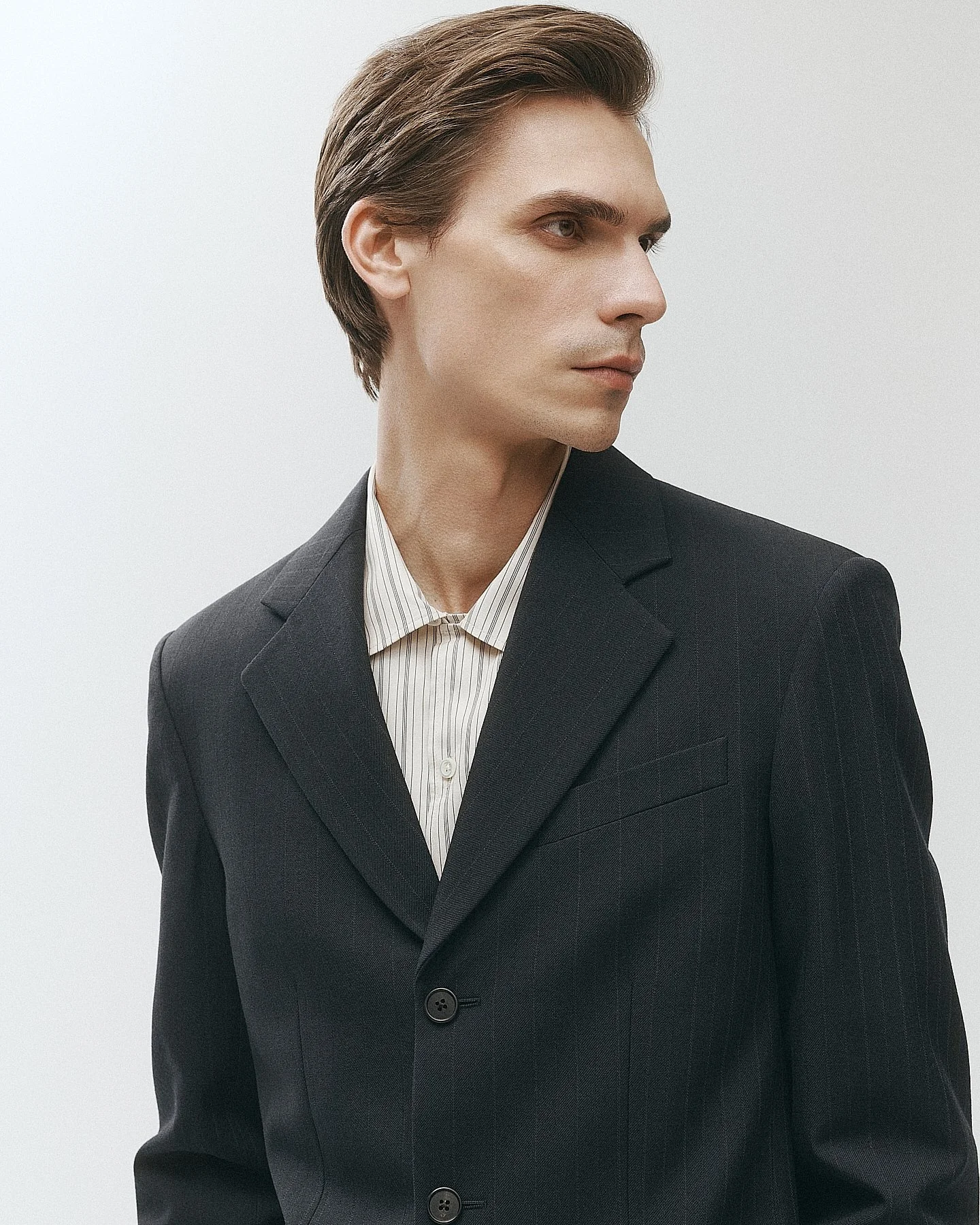 A man in a dark pinstripe suit with a light-colored striped shirt, looking to the side against a plain white background.