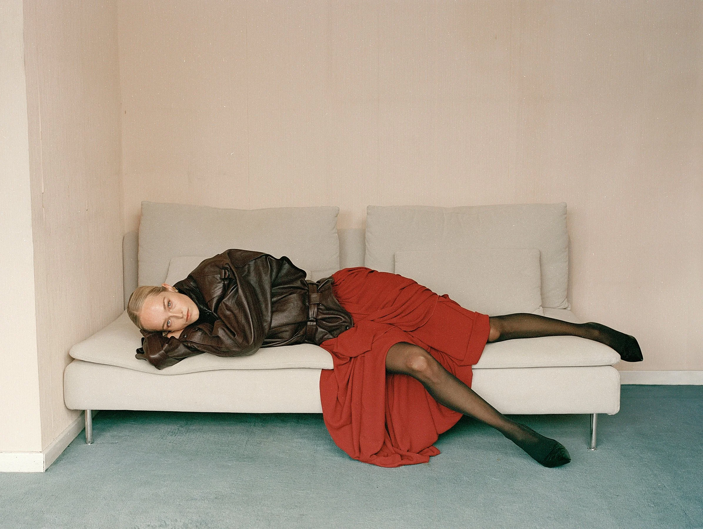 A woman lying on a white sofa in a room with light-colored walls and teal carpet. She is wearing a black leather jacket, a red skirt, black stockings, and black shoes, with blond hair styled back, looking at the camera.