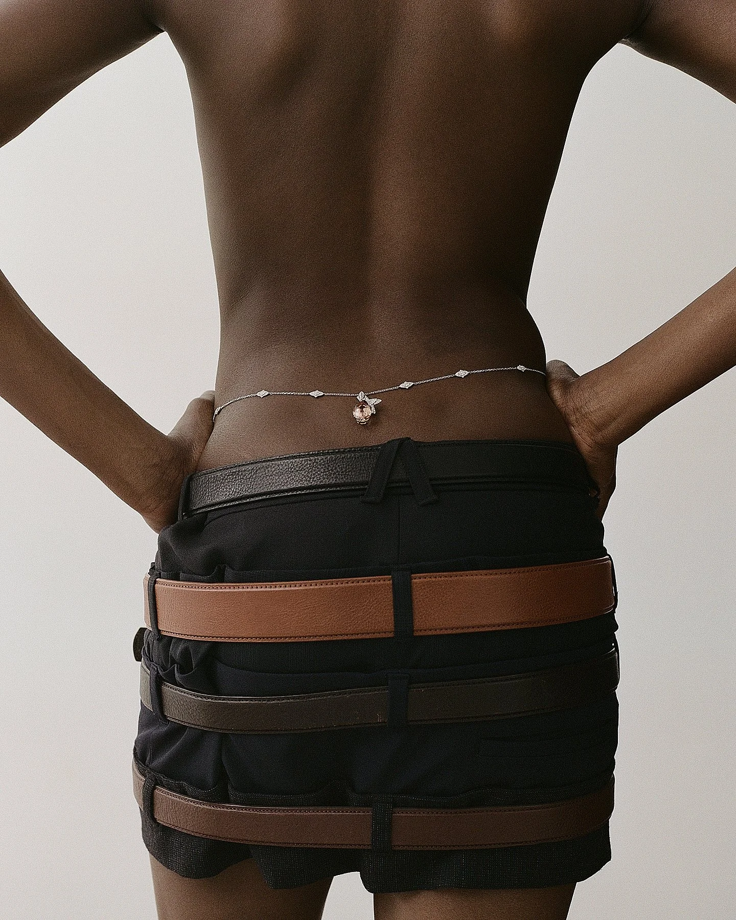 Close-up of a person's torso, wearing layered black shorts with brown and black belts, and a necklace with a large pink gemstone pendant.