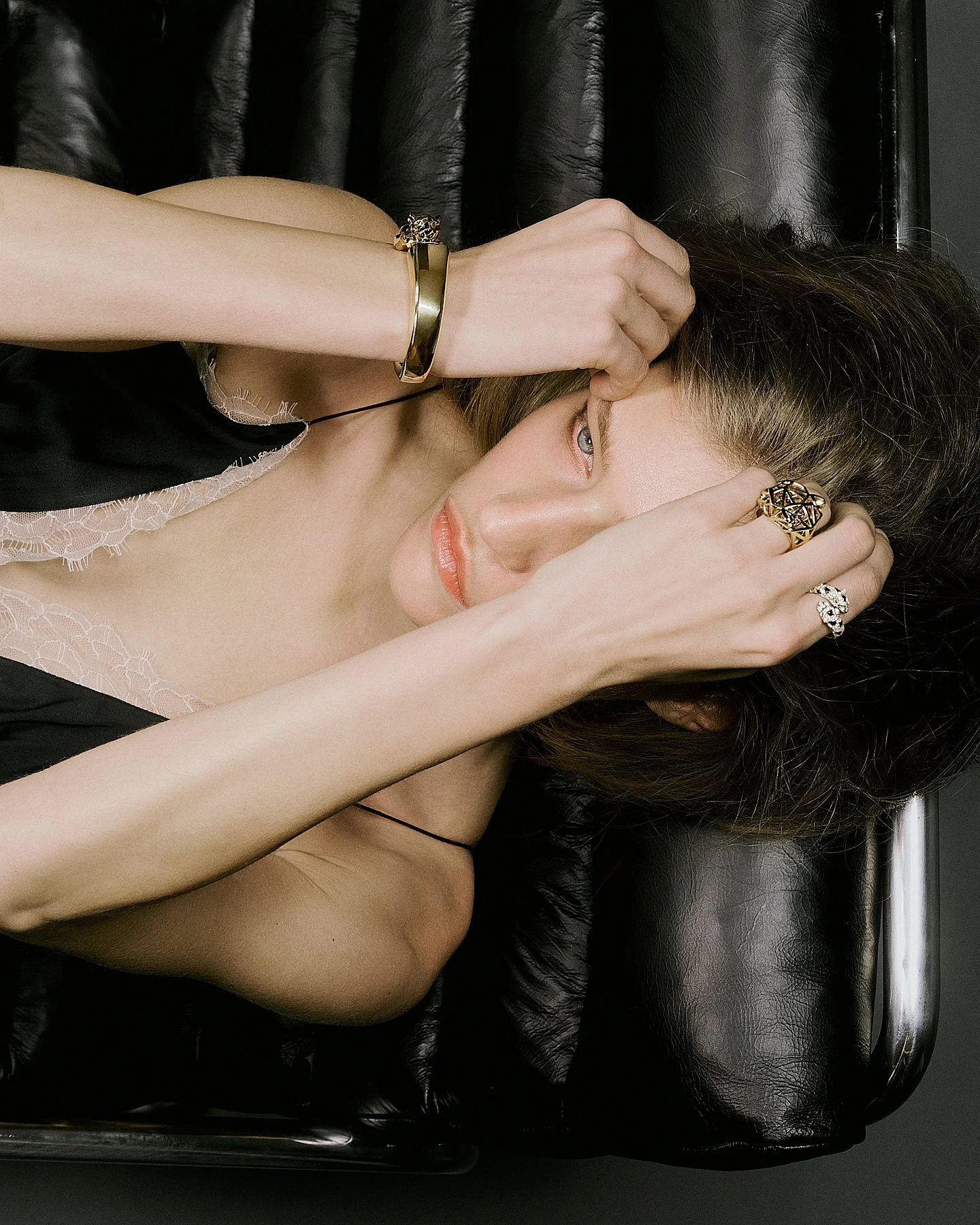 A woman with fair skin and brown hair lying on a black leather surface, wearing jewelry including a gold bracelet, a large gold ring, and a silver ring with diamonds. She is touching her face and has a black lace-trimmed garment visible.