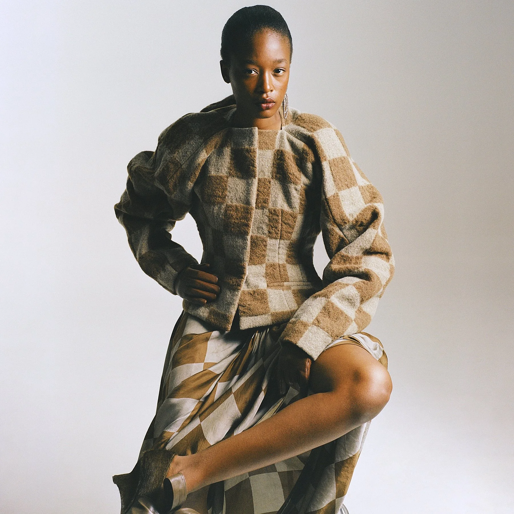 A woman with dark skin and short hair, wearing a checkered brown and beige jacket, sitting with one knee up and looking at the camera against a plain light gray background.