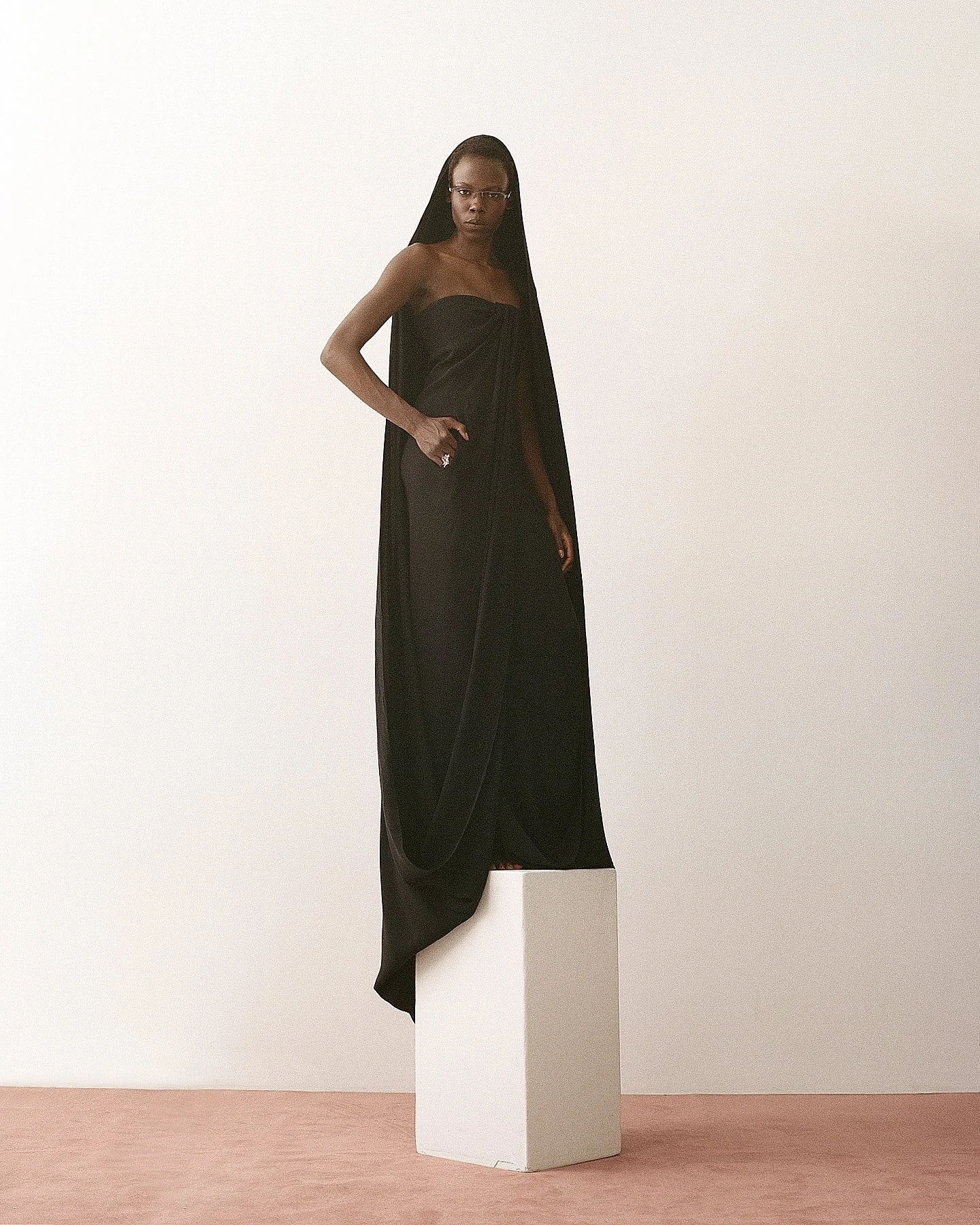 A woman standing on a white pedestal, wearing a long black dress with a sheer overlay, glasses, and dark hair, against a simple light-colored background.
