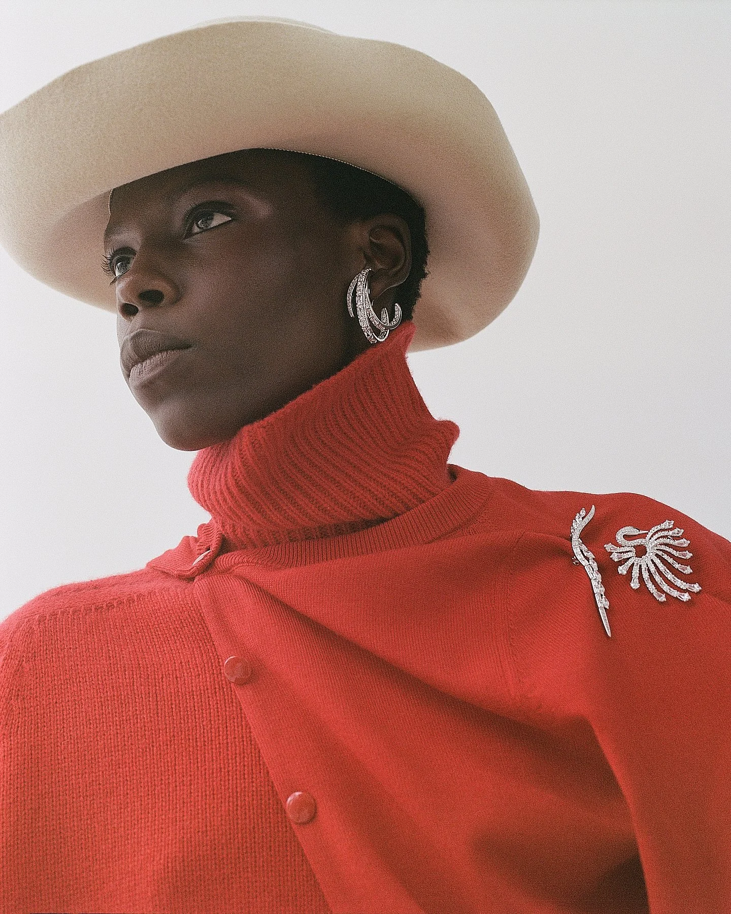 A woman with dark skin wearing a large beige hat, a red turtleneck sweater, and large silver earrings. The sweater has decorative embellishments on the shoulder.