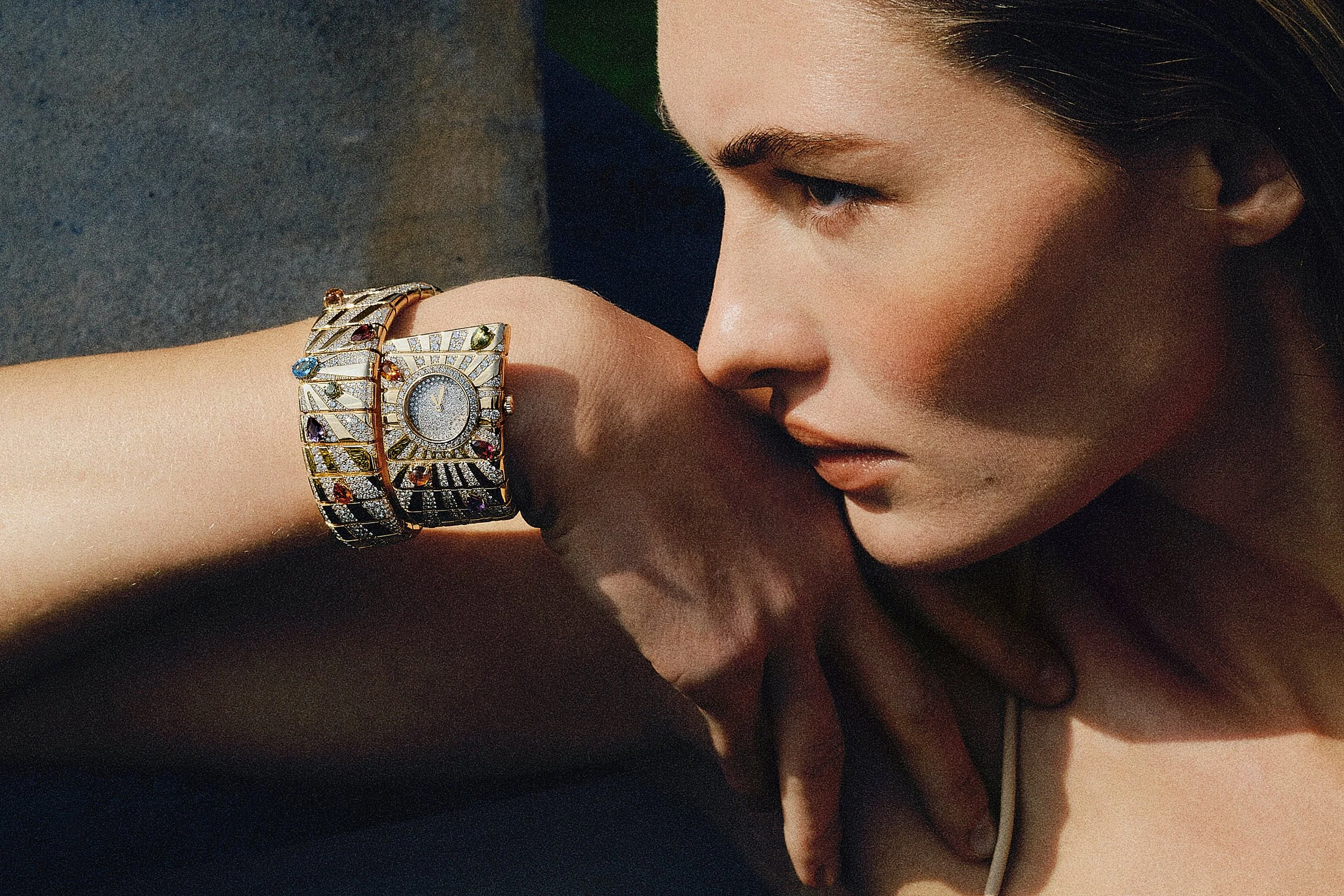 Close-up of a woman's face looking at her wristwatch, with dark brown hair, smooth skin, and wearing a gold and diamond bracelet watch.
