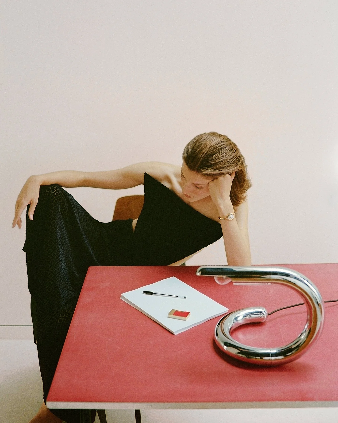 A woman with shoulder-length hair in a black off-the-shoulder dress leans on a red table, resting her head on her hand, with a notebook, pen, and eraser on the table. A metal faucet is in the foreground.