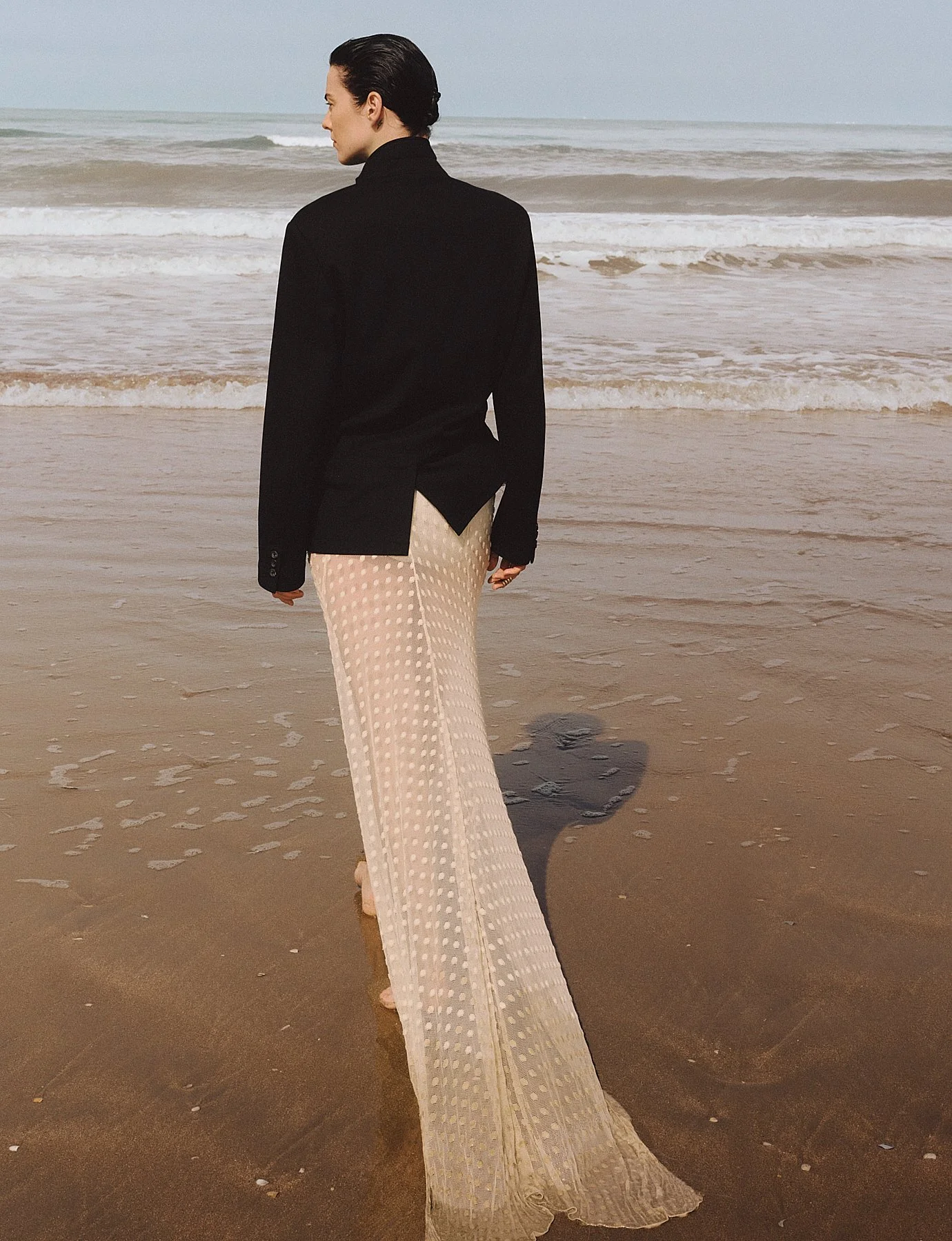 Woman standing on the beach facing the ocean, wearing a black jacket and wide-legged beige patterned pants, with wet hair and casting a shadow on the sand.