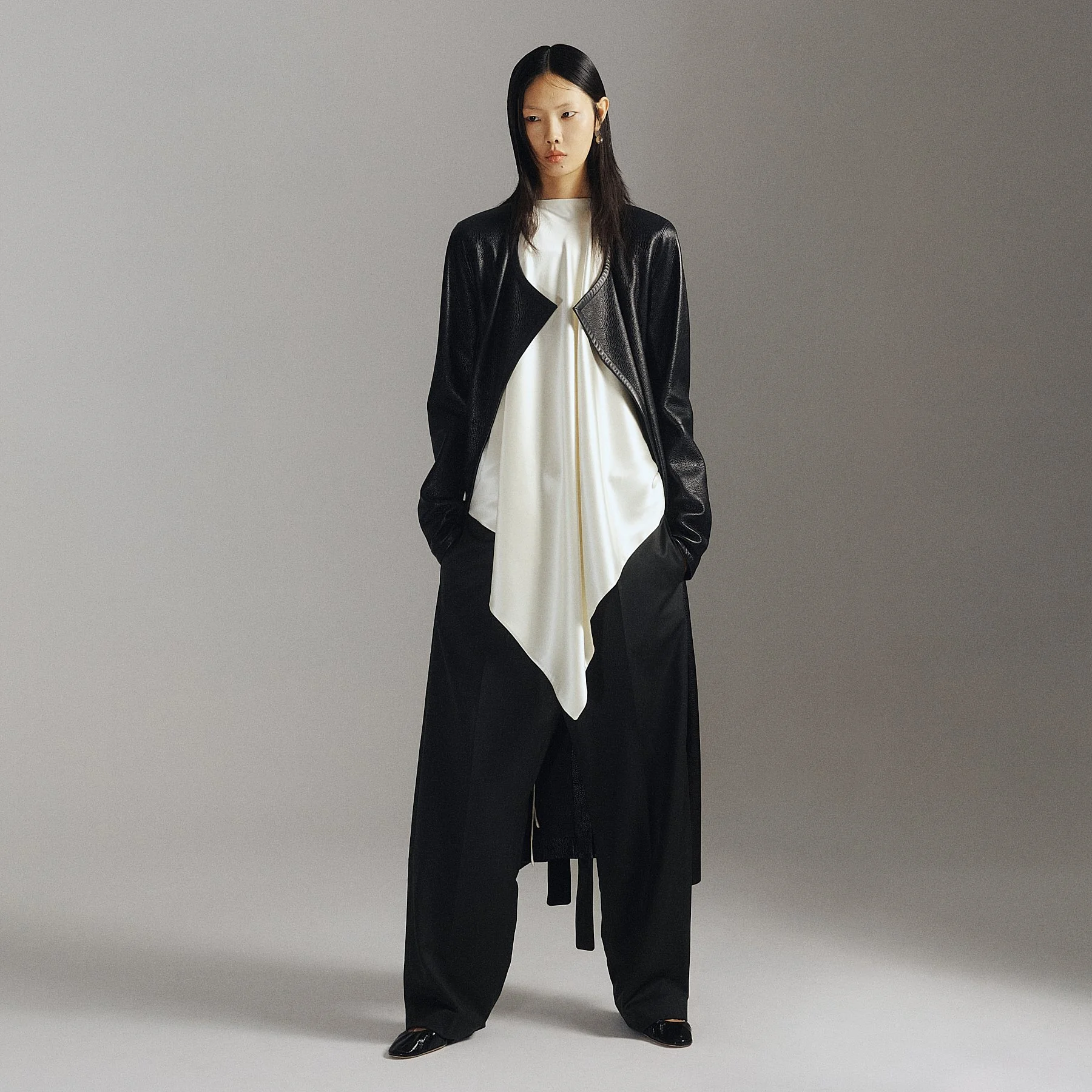 A woman with long black hair wearing a black leather jacket over a white dress with black wide-leg pants, standing against a plain gray background.