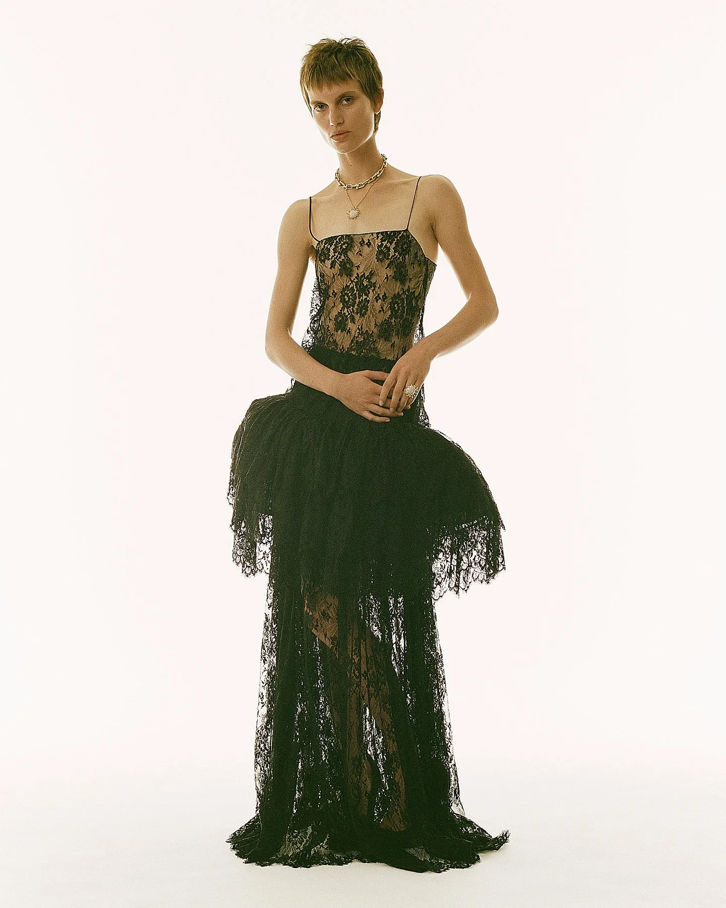 A woman with short hair wearing a black lace dress with spaghetti straps, accessorized with layered necklaces and rings, standing against a plain white background.