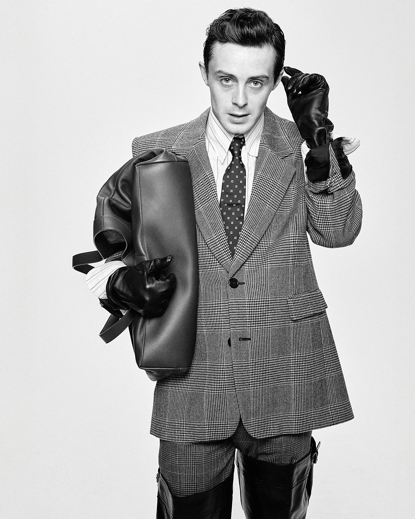 Black-and-white portrait of a man wearing a plaid suit and tie, holding a backpack, and adjusting his earring, with black leather gloves and pants.