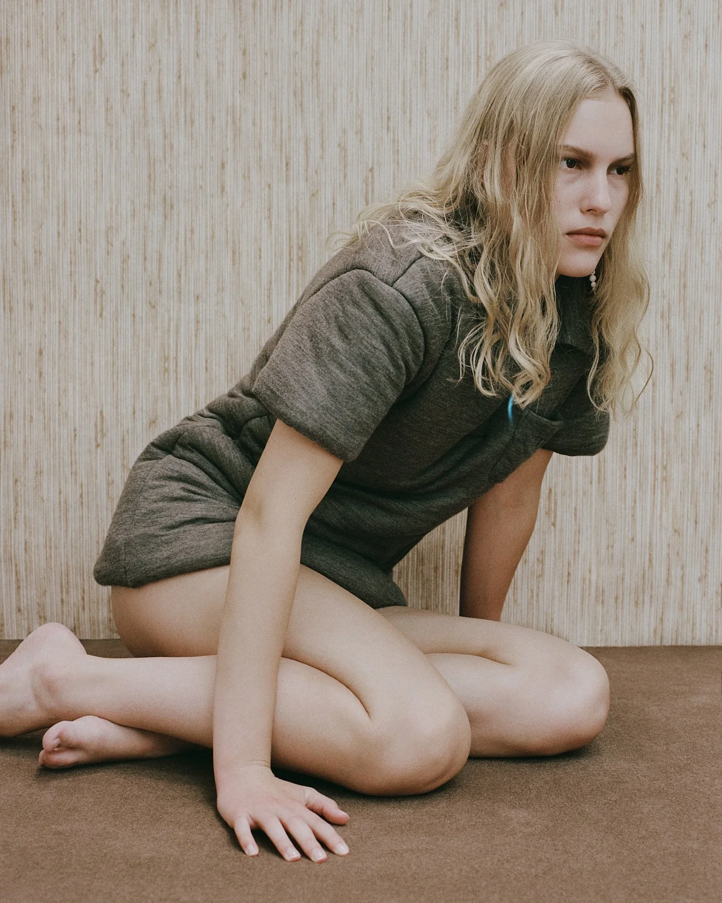 A woman with long, wavy blonde hair and fair skin in a gray dress kneeling on a brown carpeted floor in front of a beige textured wall.