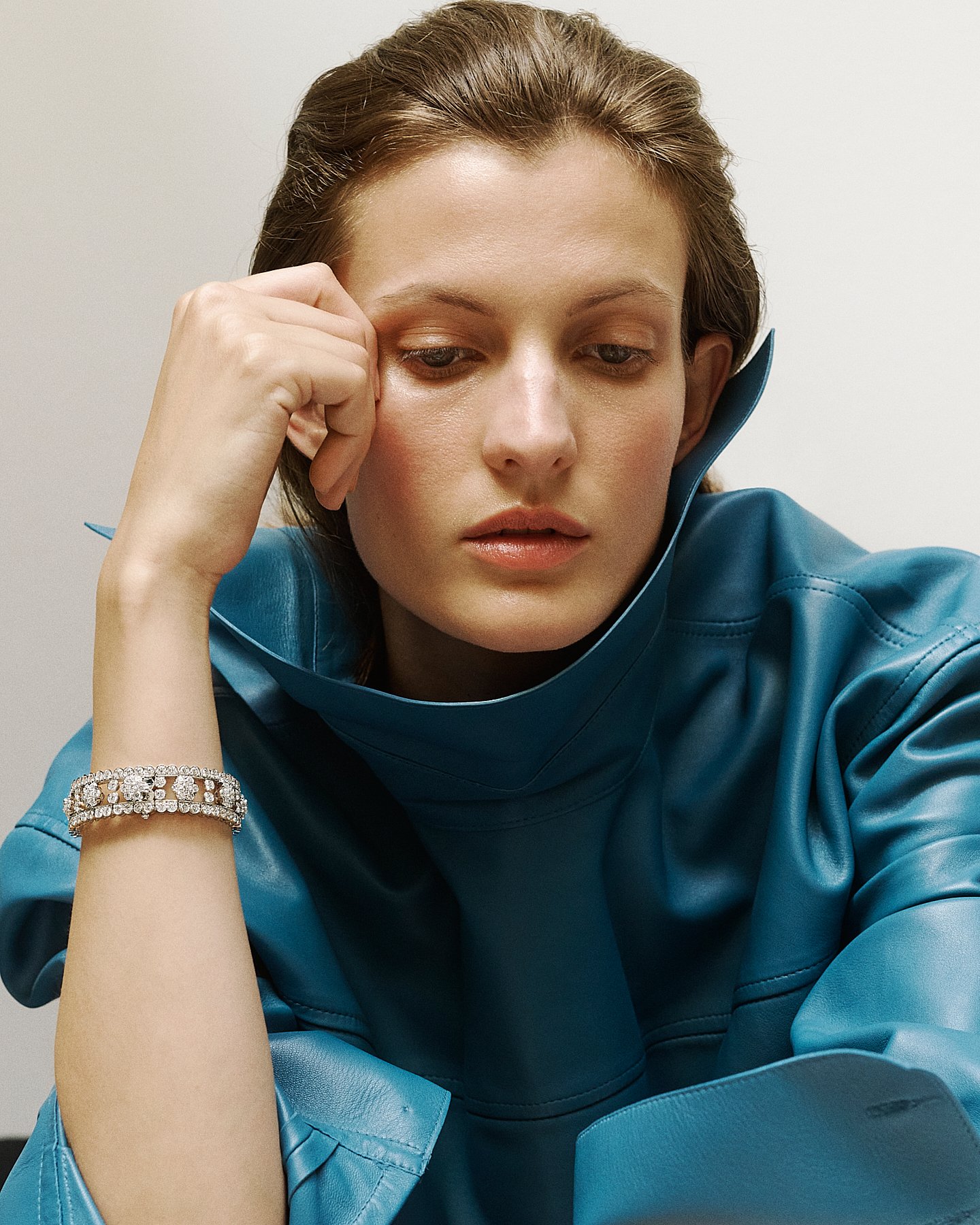 A woman with brown hair wearing a blue leather jacket and a rhinestone bracelet, resting her head on her hand, looking down.