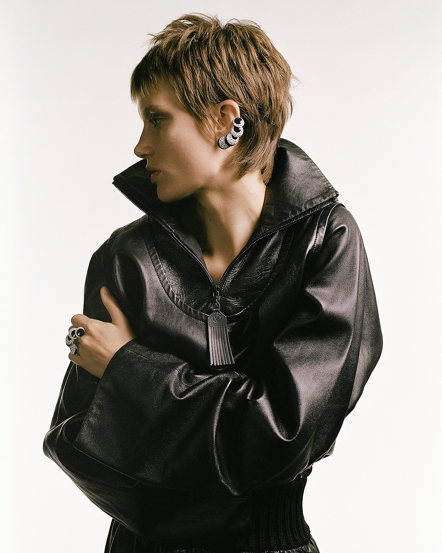 A woman with short brown hair wearing a black leather jacket, large sparkling earrings, a matching ring, and a large ring with a black and gold design, standing against a plain white background.
