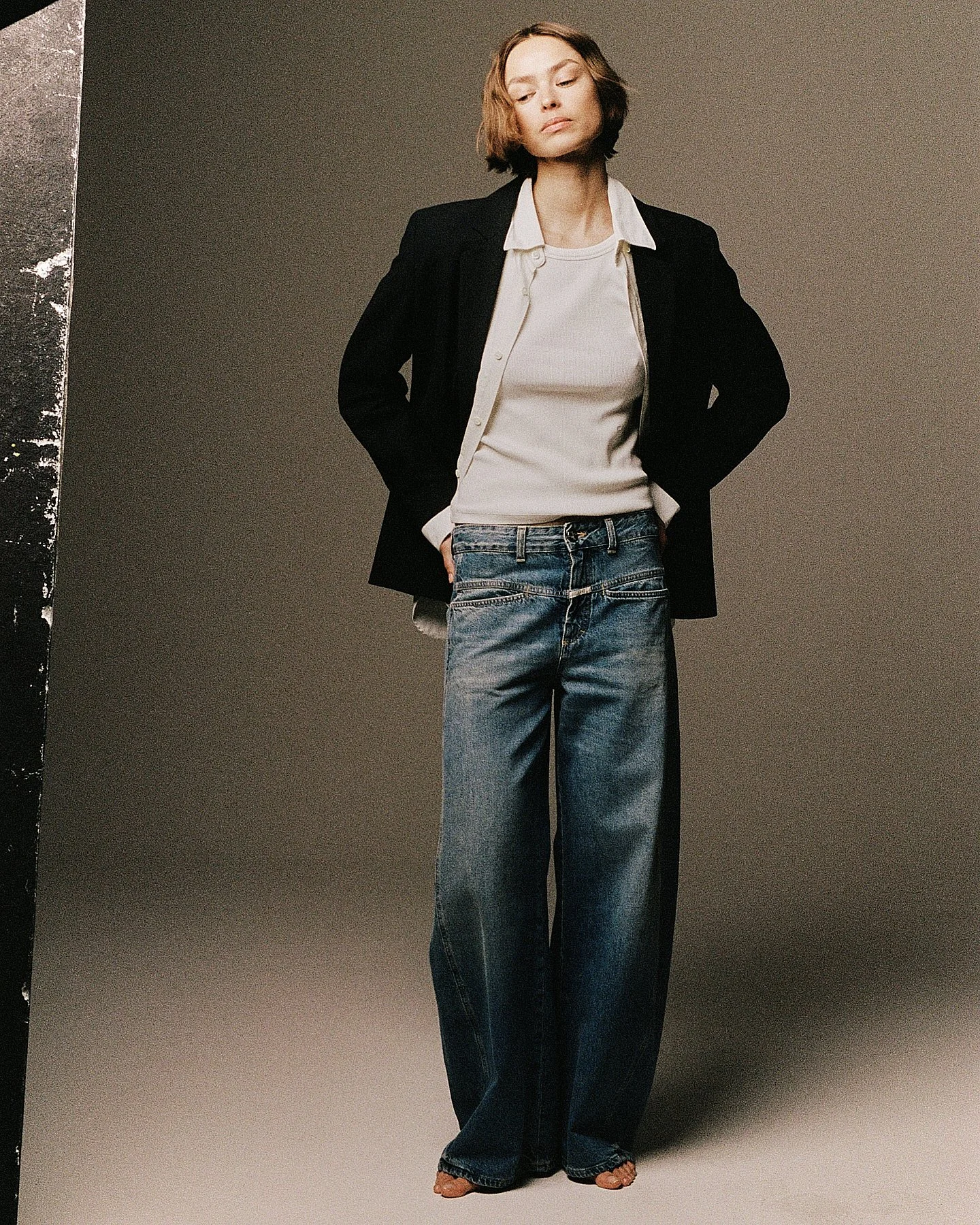 Woman with short hair wearing a white shirt, black blazer, and loose-fitting blue jeans, standing against a plain background.